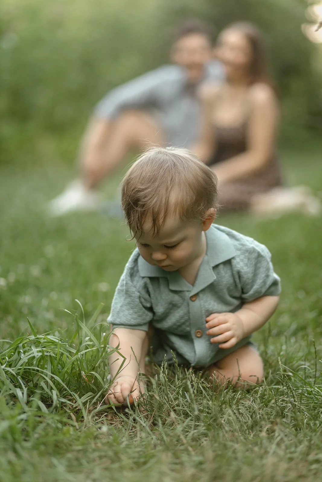 A young child crawling on the grass with two adults sitting in the blurry background at Lincoln Park.