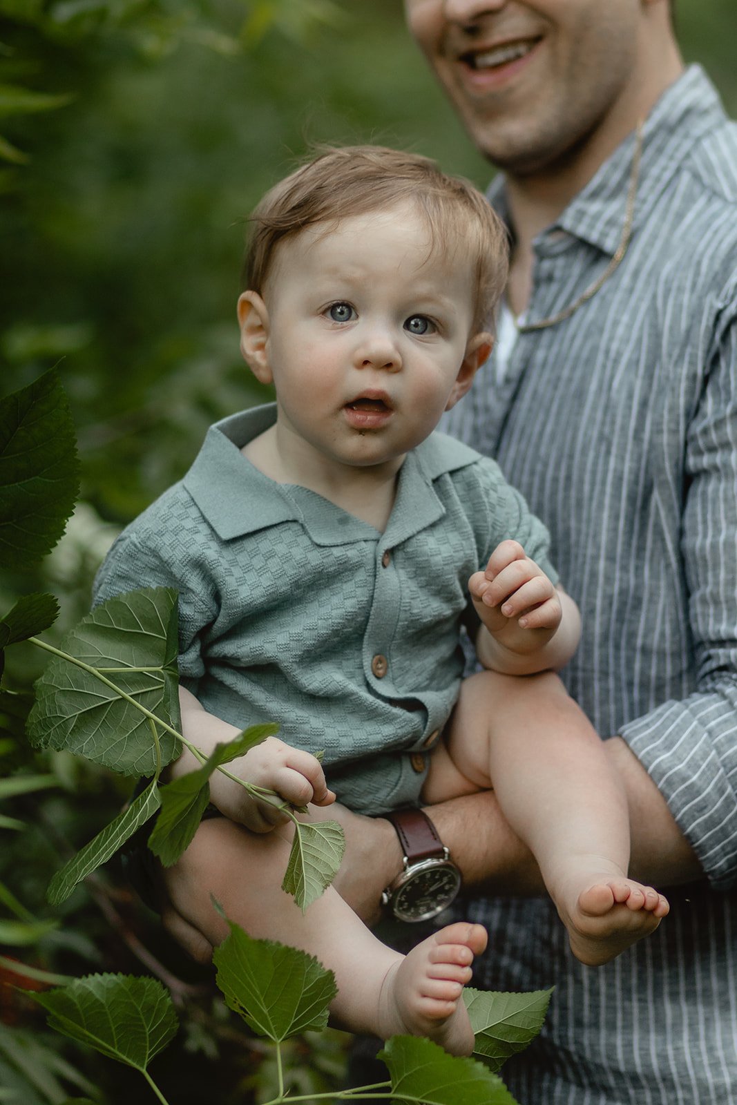 A young child with blue eyes and tousled light brown hair sitting on an adult's lap outdoors, surrounded by green leaves, with a surprised expression on his face.