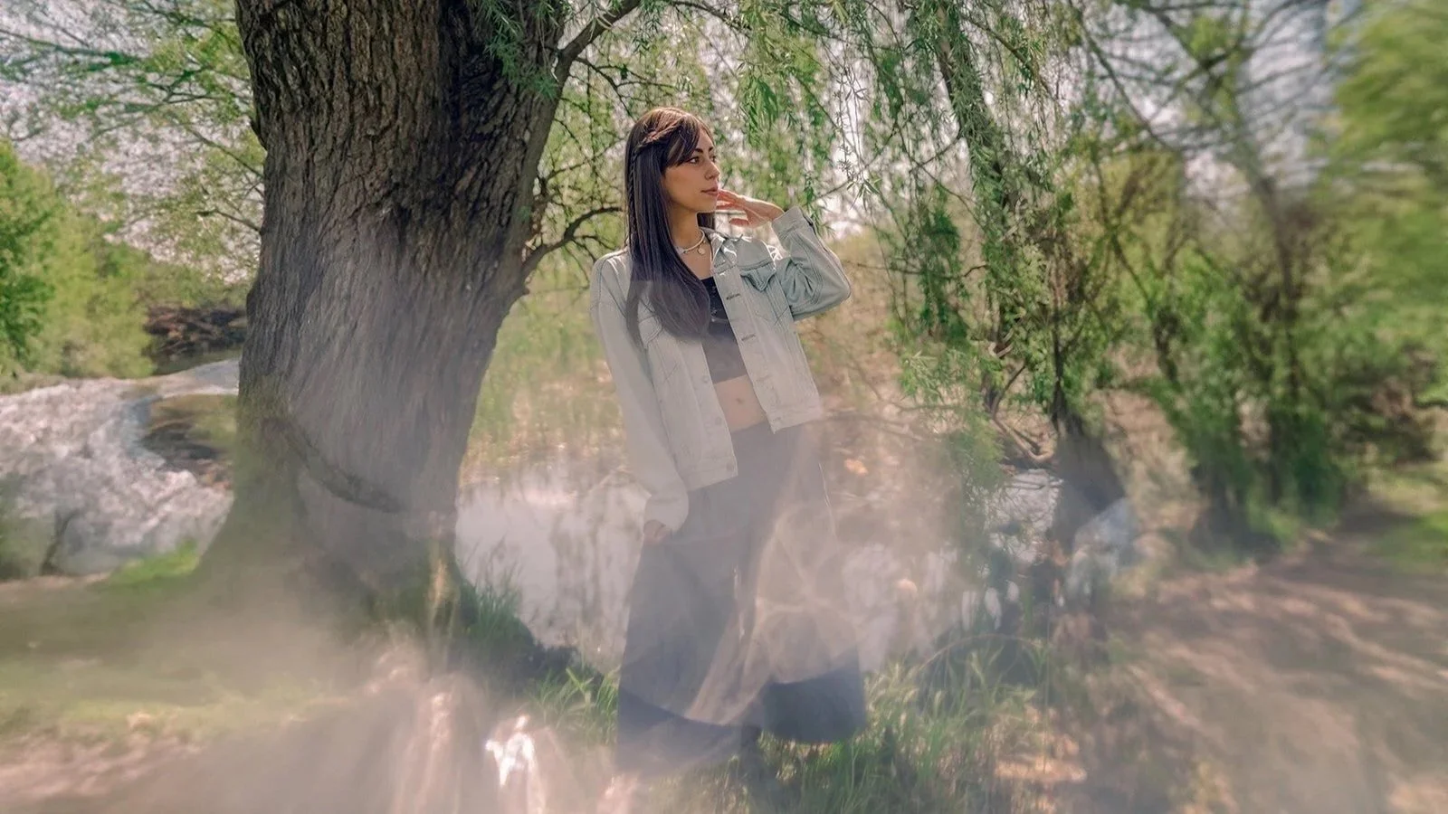 A young woman stands next to a large tree in a lush, green outdoor setting during daytime, with a river or stream in the background.