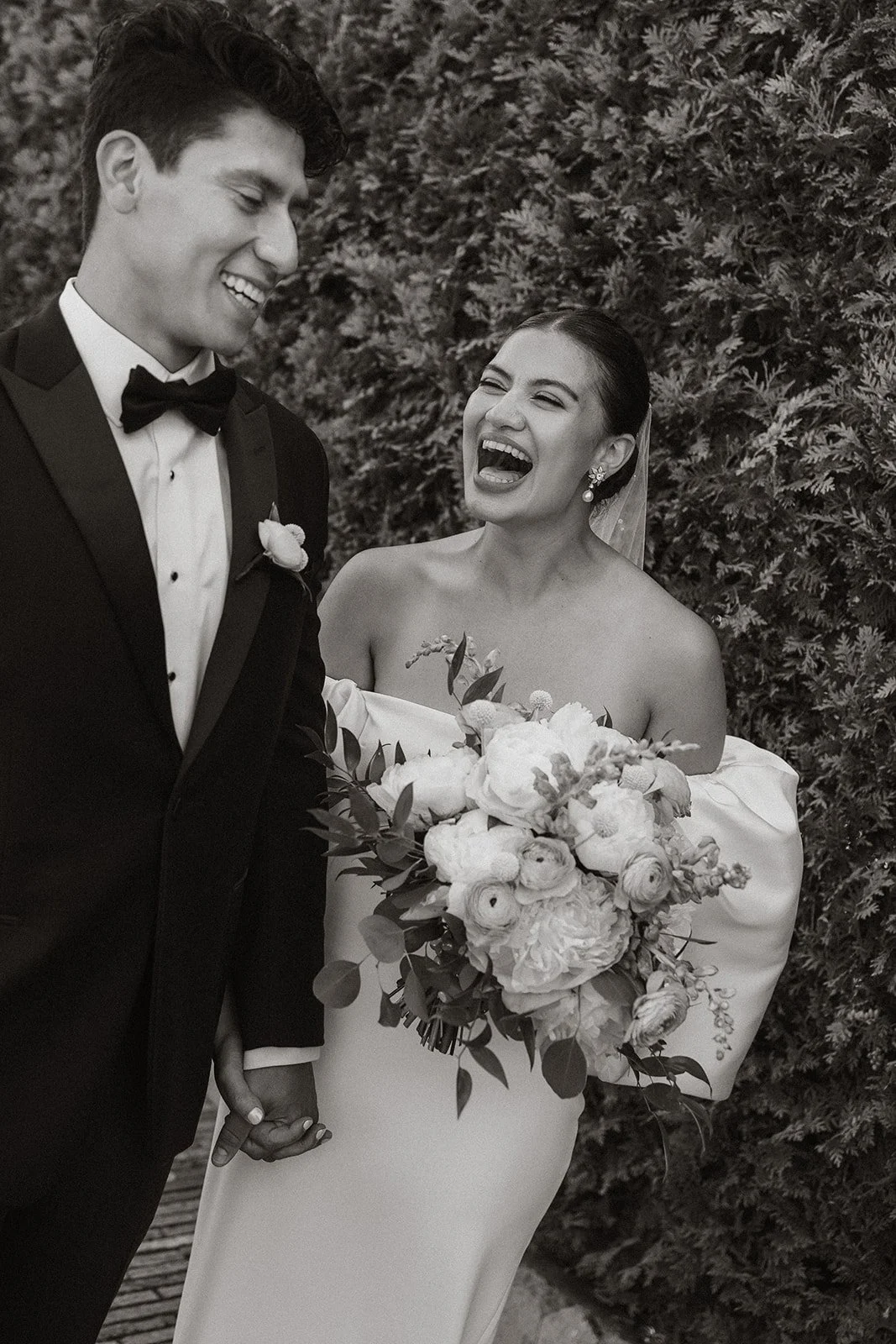 A black and white photo of a bride and groom at their wedding. The groom is wearing a tuxedo with a bow tie, smiling, while the bride, holding a large bouquet of flowers, is happily laughing and looking at him. They are holding hands and standing in front of a bushy background.