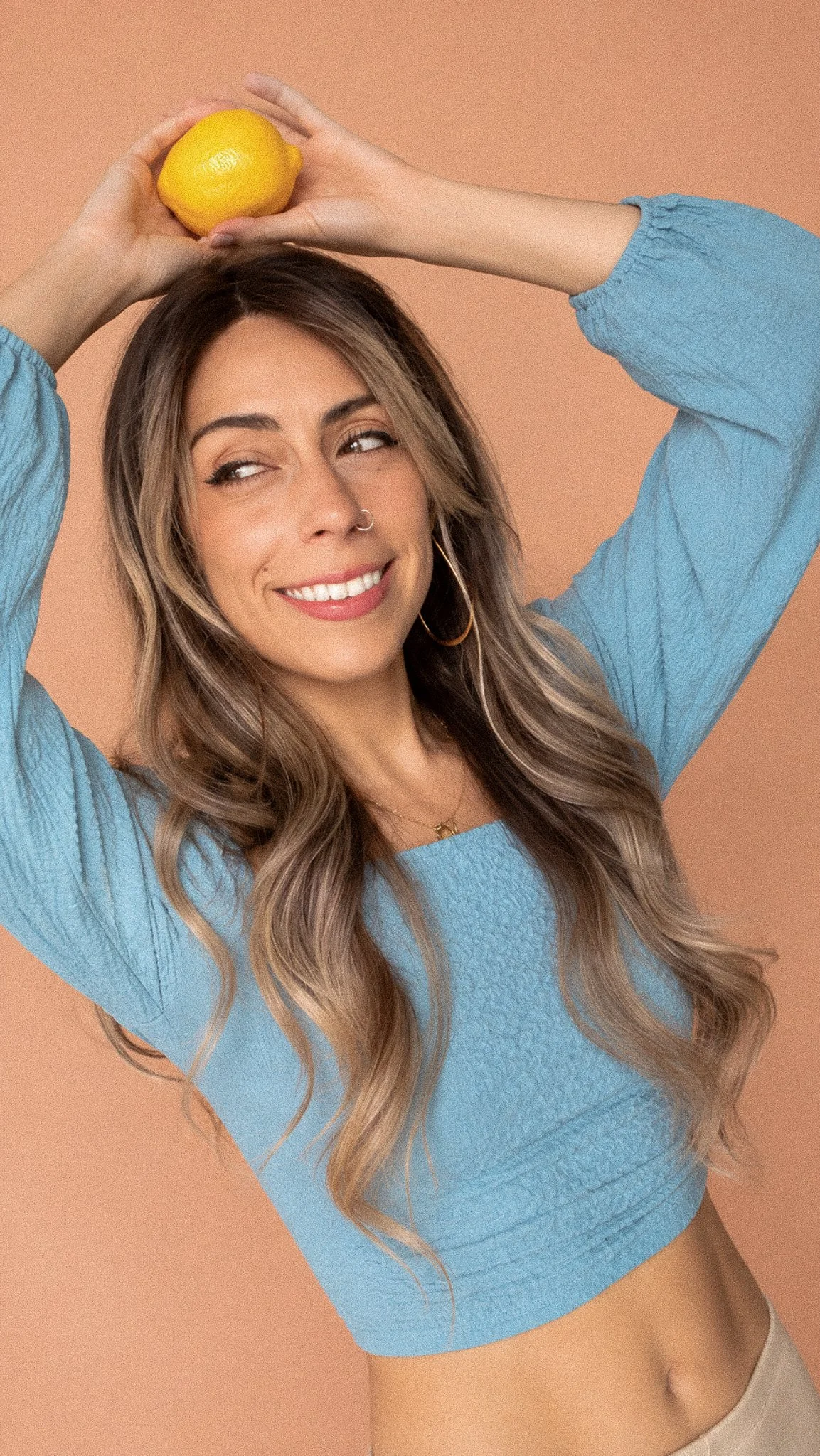 A smiling woman with long wavy hair, wearing a blue top, holding a lemon above her head against a peach background.