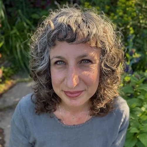 Person with curly hair wearing a gray shirt against a green botanical background.