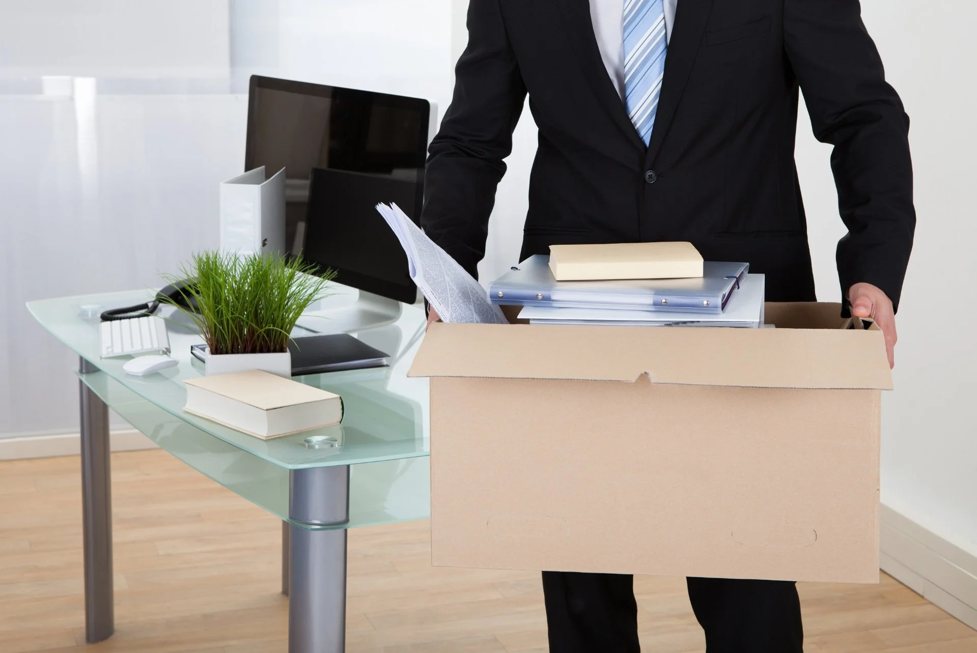 Person in suit moving a cardboard box filled with files and papers in an office, with a desk, computer, and office supplies in the background.