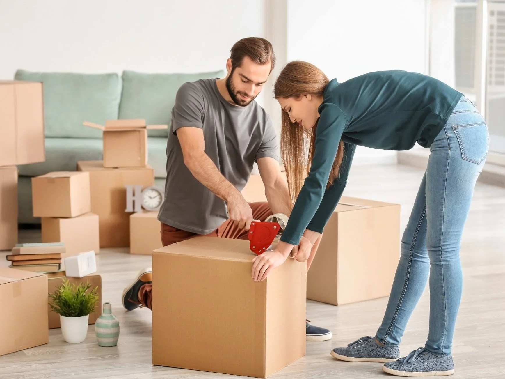 A young woman and man are packing moving boxes with packing tape in a bright living room.