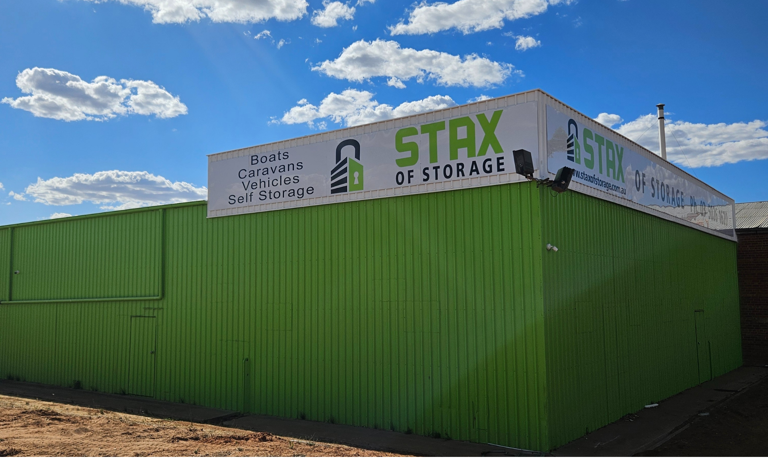 Green building with a white sign reading 'Stax of Storage' and listing items like boats, caravans, vehicles, and self-storage. Blue sky with clouds in the background.