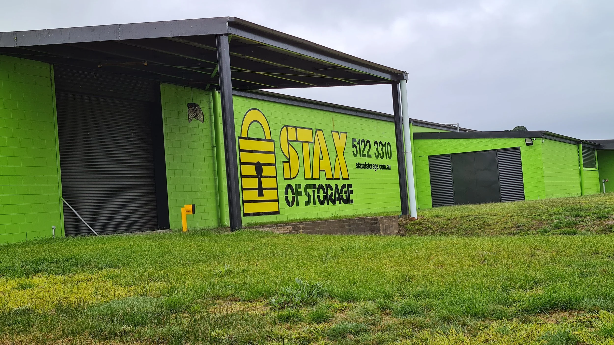 Green building with a sign reading 'Stax of Storage' and a phone number, with black roller doors and a grassy foreground.