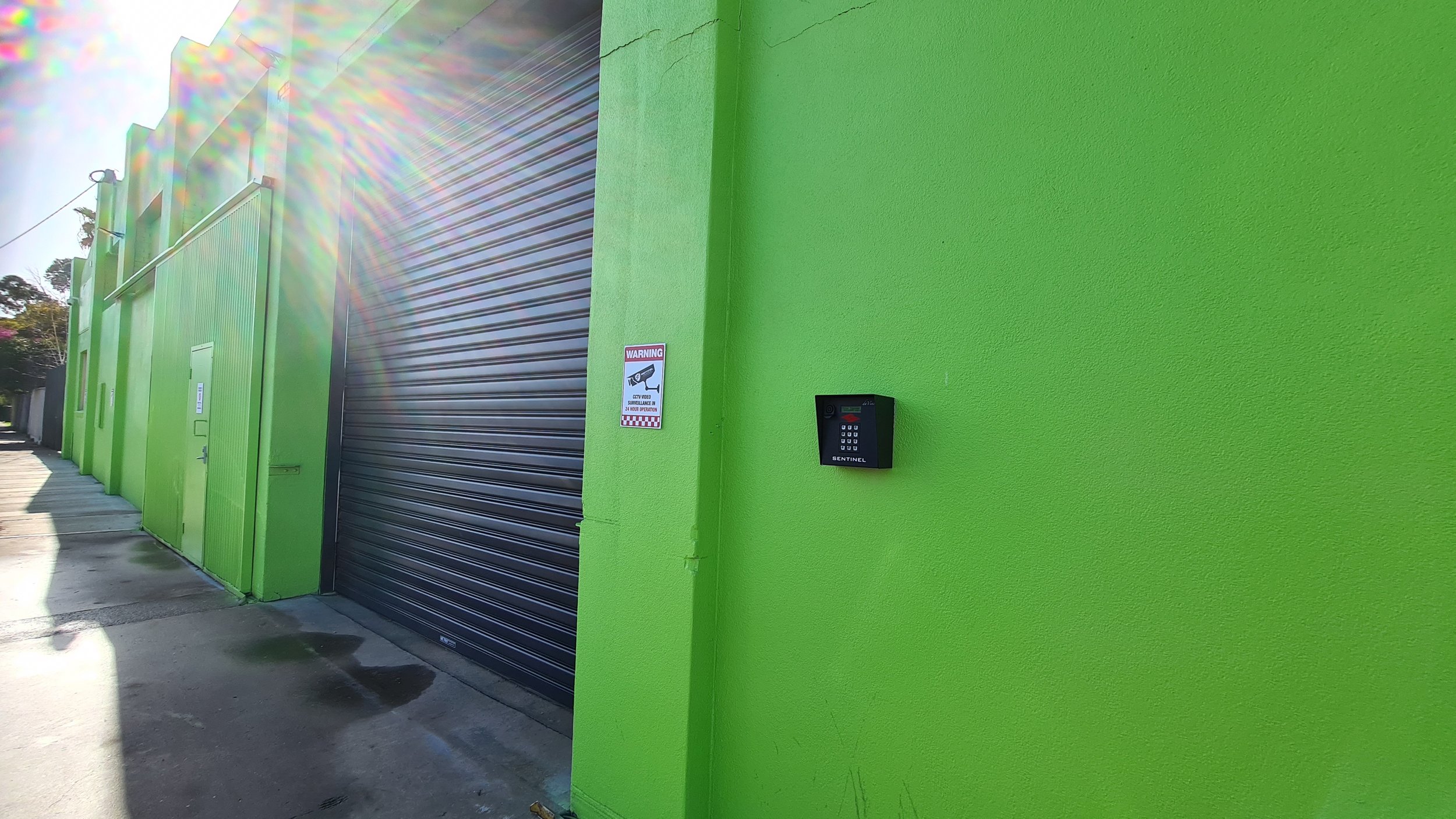 Bright green commercial building with a black security keypad on the wall and a warning sign about surveillance cameras, sidewalk in front, with the sun shining brightly creating a lens flare.
