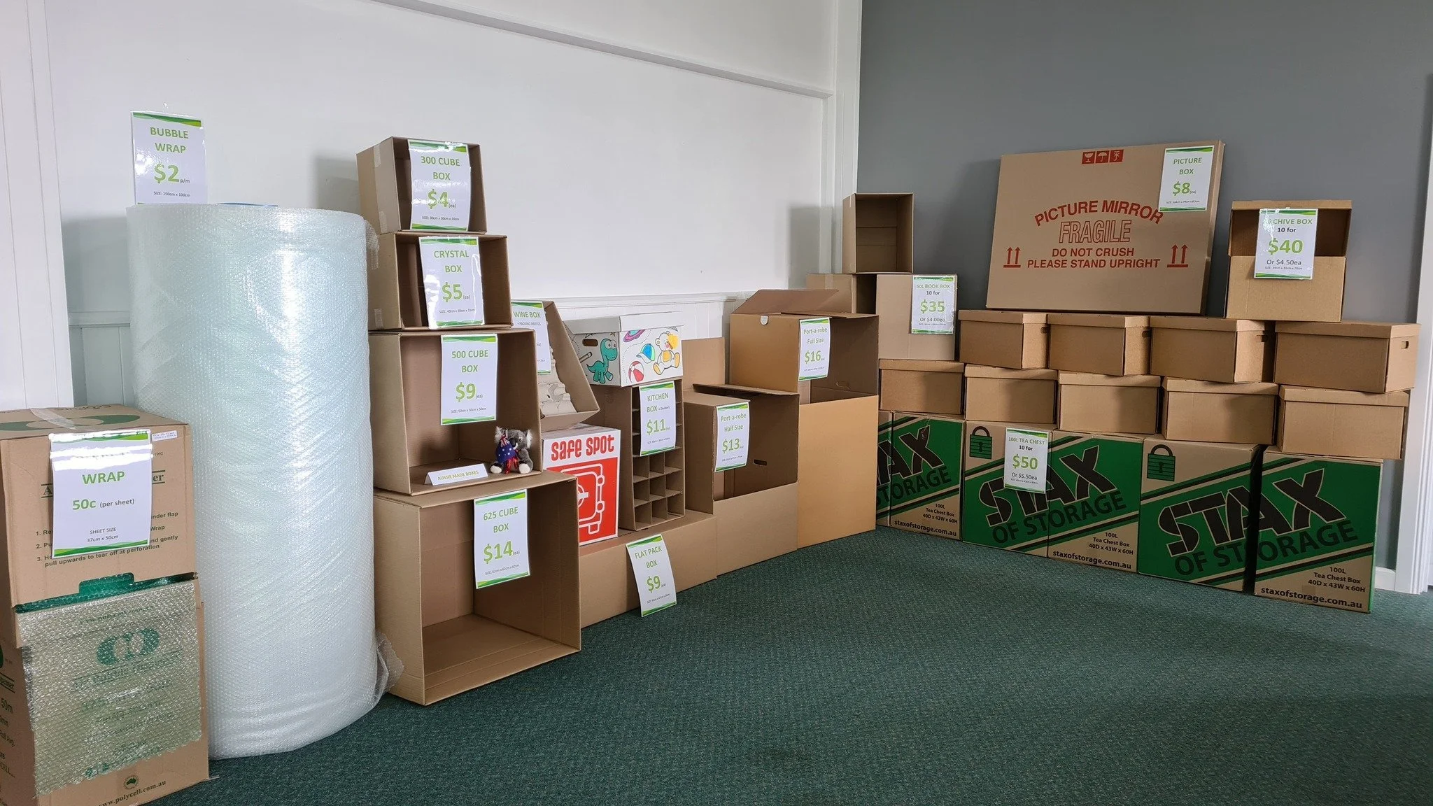 Room filled with various cardboard boxes, some labeled with prices and sizes, arranged against beige and gray walls, some stacked on top of each other, with a large roll of bubble wrap on the left side and green storage boxes at the bottom right.
