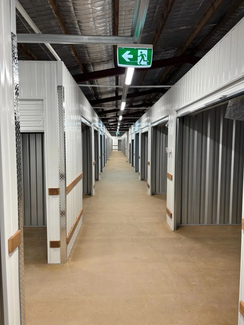 A long corridor with storage units on both sides, metal doors, and an emergency exit sign hanging from the ceiling showing an arrow pointing left and a running figure.
