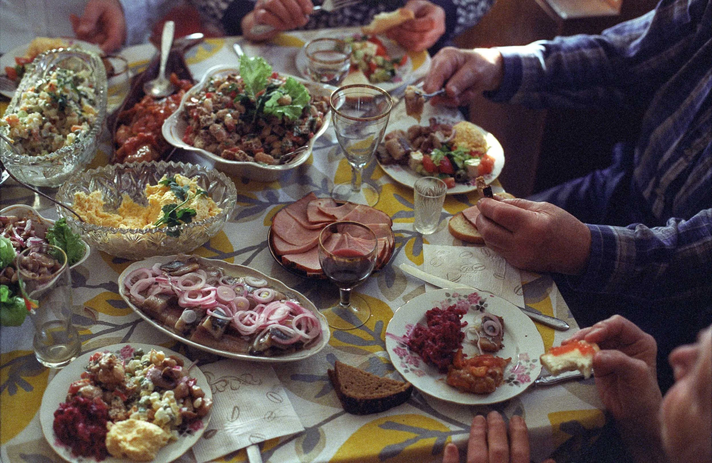 A documentary photograph of a family gathered around a table filled with various dishes, including salads, meats, potatoes, bread, and wine, enjoying a meal together.