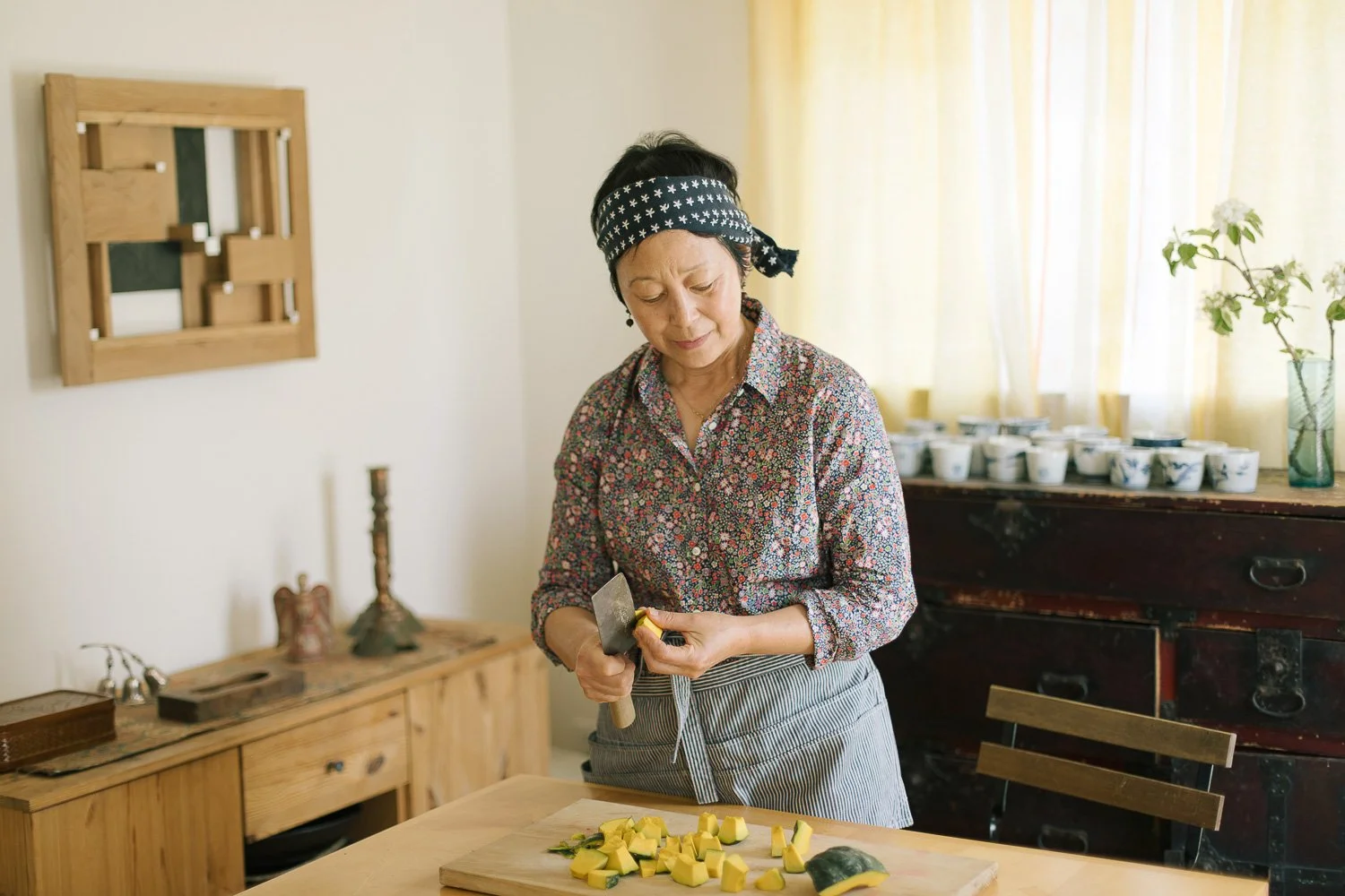 An elderly woman with a floral blouse and a headscarf chopping yellow squash on a wooden cutting board in a cozy kitchen.