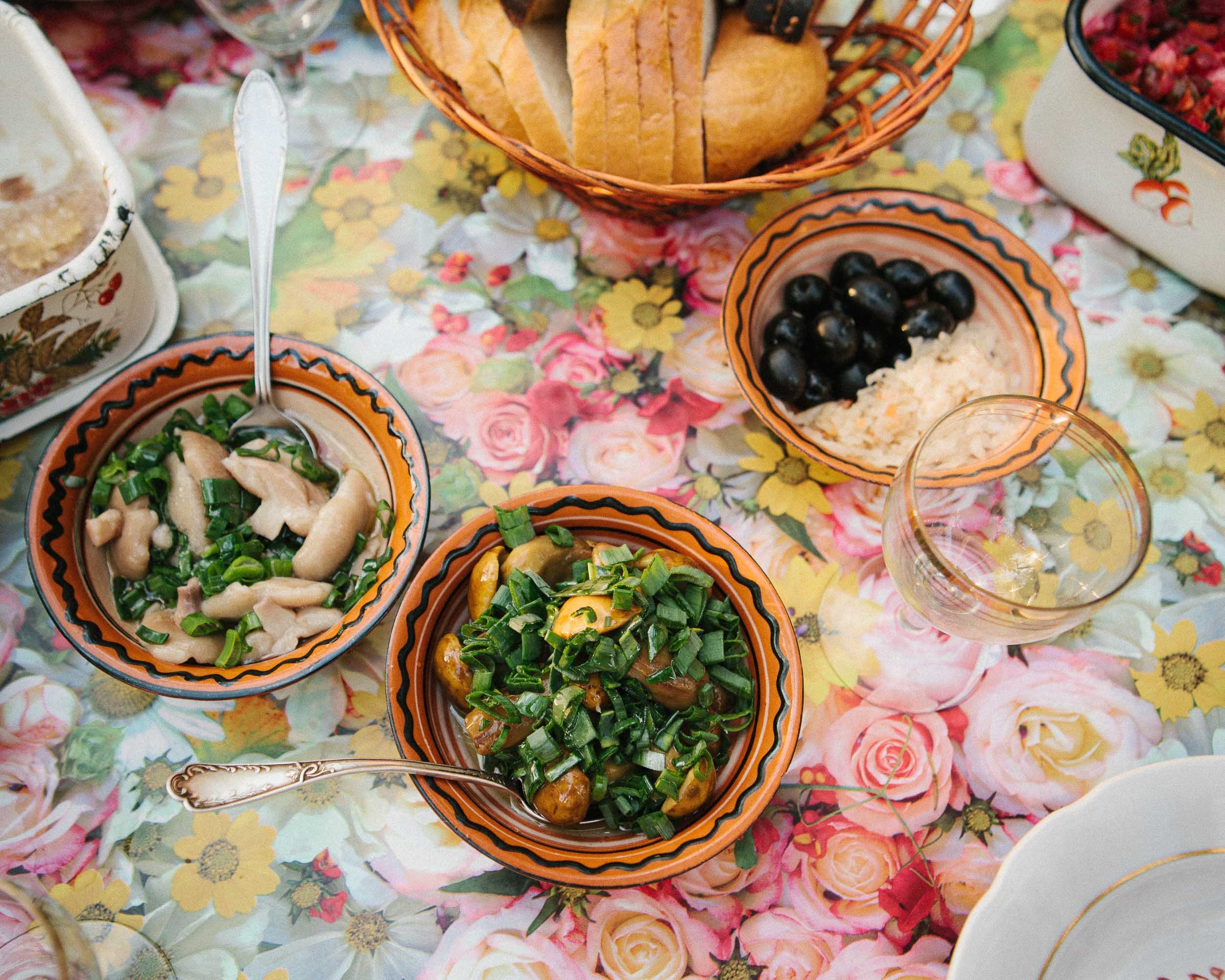 An overhead lifestyle photograph of a table set for a meal with bowls of salad topped with green onions, a bowl of rice with black olives, a basket of bread, and a glass of white wine on a floral tablecloth.