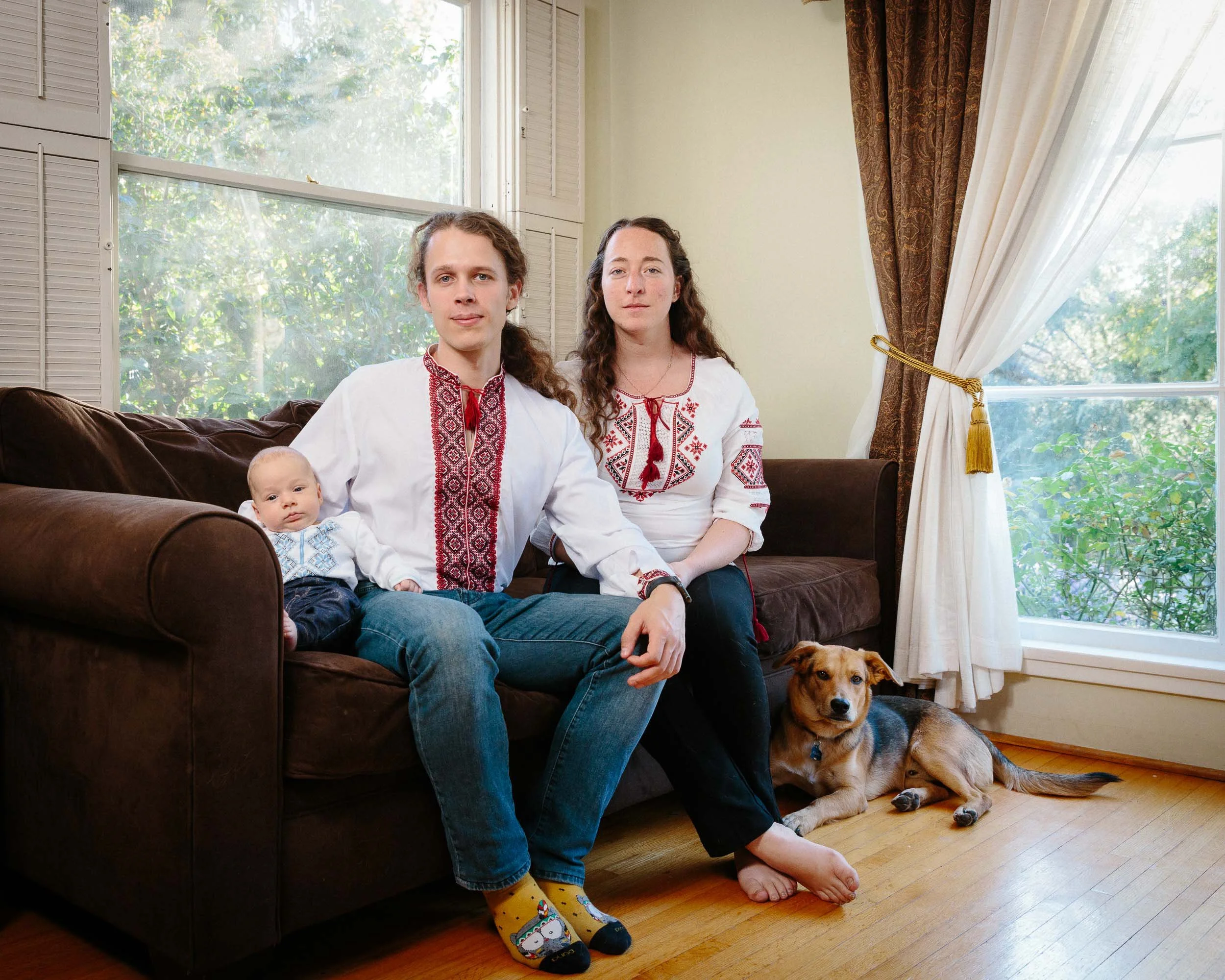An editorial environmental portrait of a family of three sitting on a brown couch in a living room with a dog lying on the floor nearby. The family is dressed in traditional embroidered clothing, with a baby and two adults.