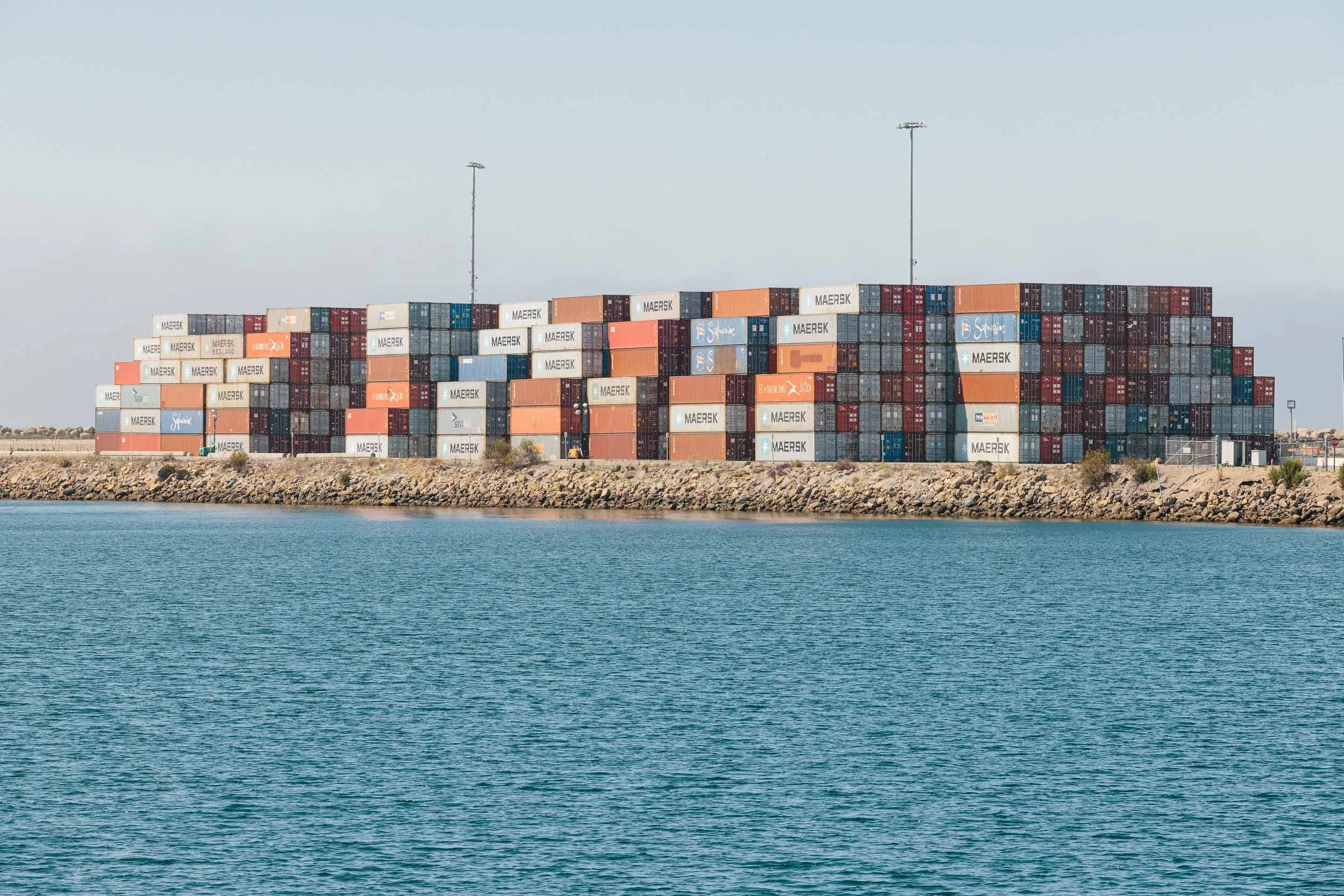 A landscape photograph of a large stack of shipping containers on a dock by the water with a clear sky in the background.