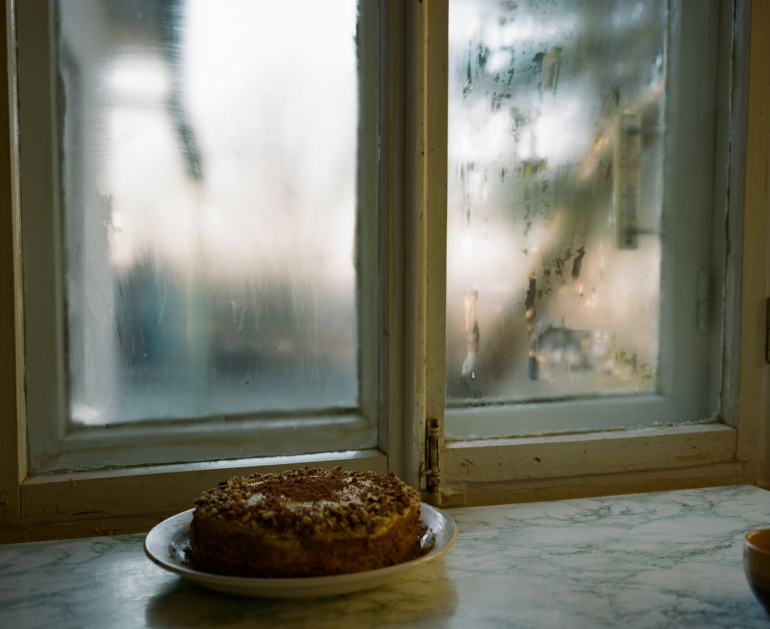 A documentary still life photograph of chocolate cake on a plate sits on a marble countertop in front of a foggy window.