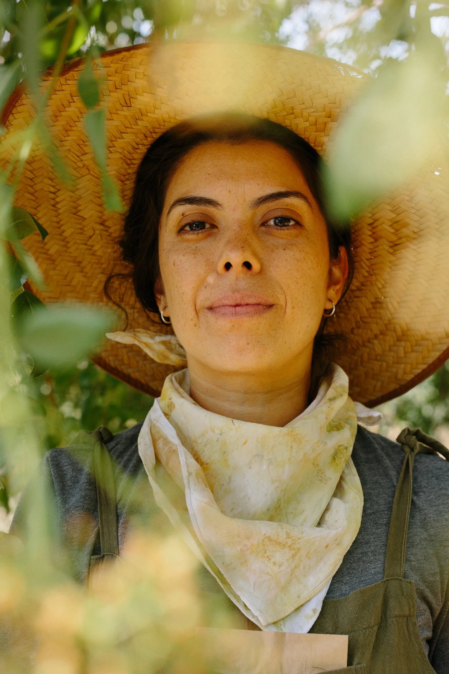 A documentary portrait of a woman wearing a wide-brimmed straw hat, a scarf, and a gray shirt, looking directly at the camera outdoors amidst greenery.
