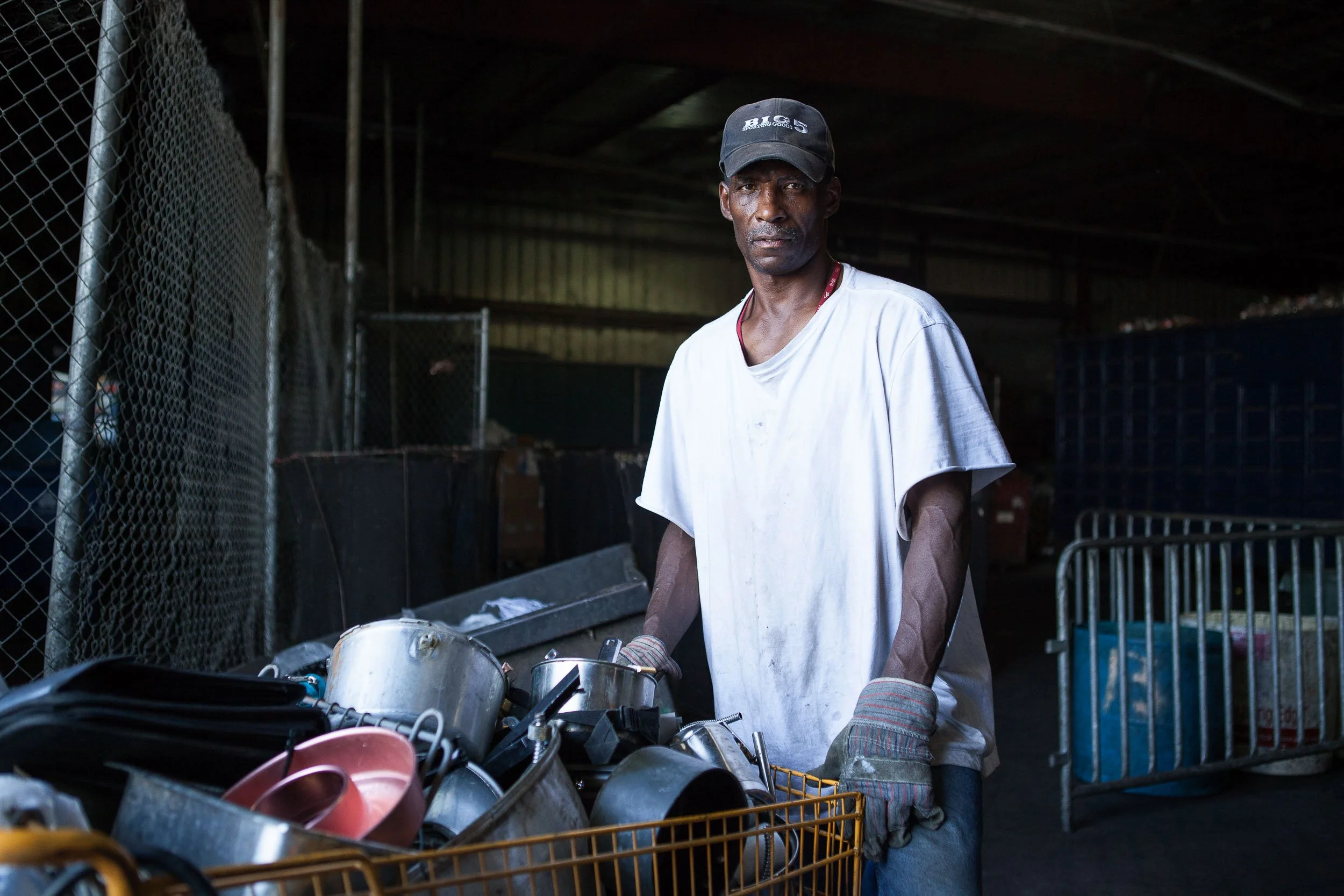 Portrait of man wearing a white t-shirt, gray work gloves, and a black cap stands behind a yellow shopping cart filled with various metal kitchen utensils. The background is a chain-link fence with storage bins inside a warehouse or industrial space.