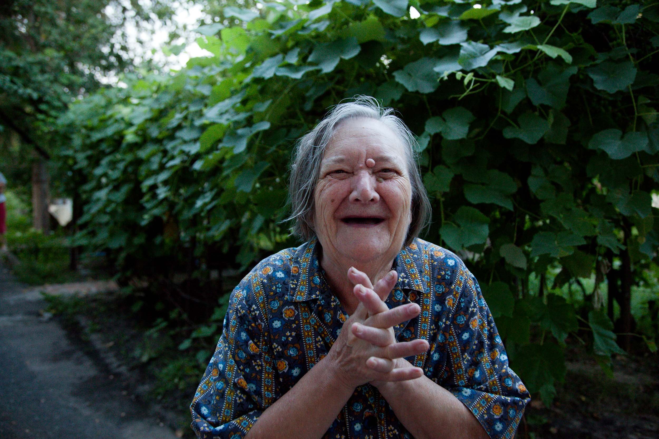 A documentary photograph of an elderly woman with gray hair and a blue patterned shirt standing outdoors in front of lush green foliage, smiling with her hands clasped.