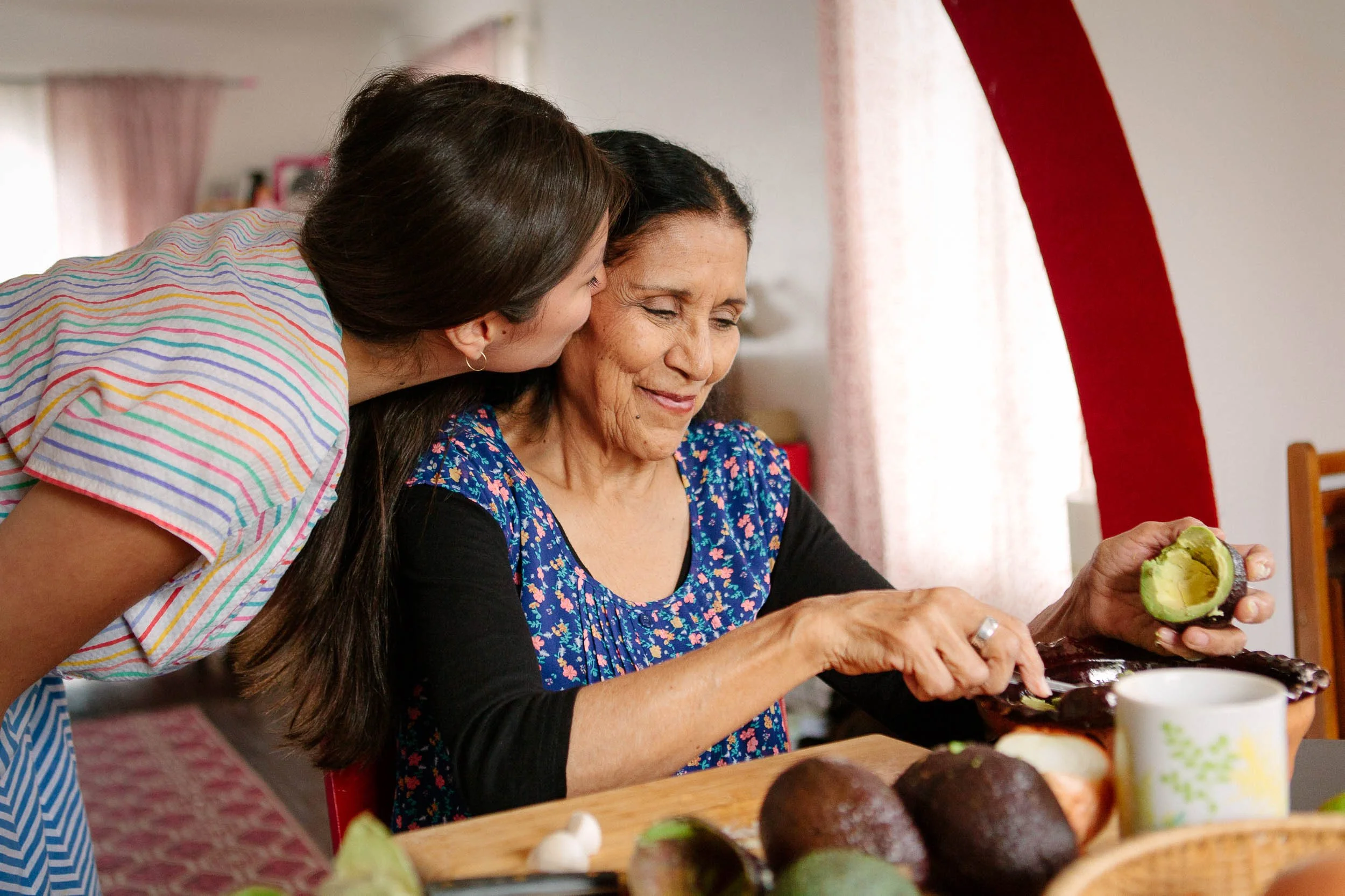 A lifestyle photograph of a young woman helping an older woman to prepare avocados at a kitchen table with avocados and a mug visible.