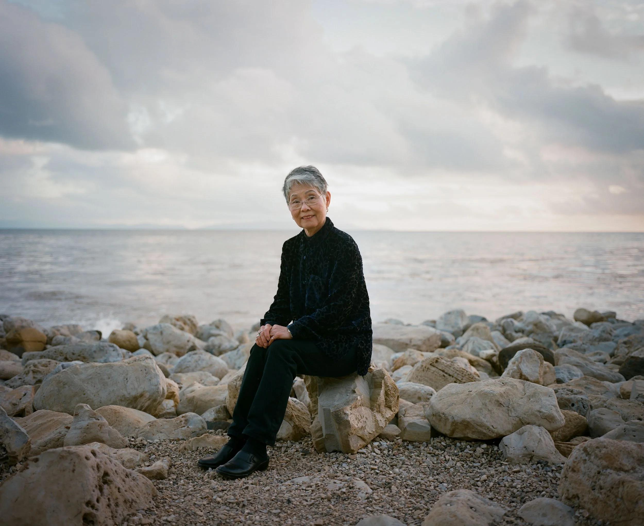 An environmental on-location portrait of a woman with short gray hair and glasses sits on a large stone at the rocky shoreline of the ocean, smiling, wearing a black jacket and black pants, with the ocean and cloudy sky in the background.
