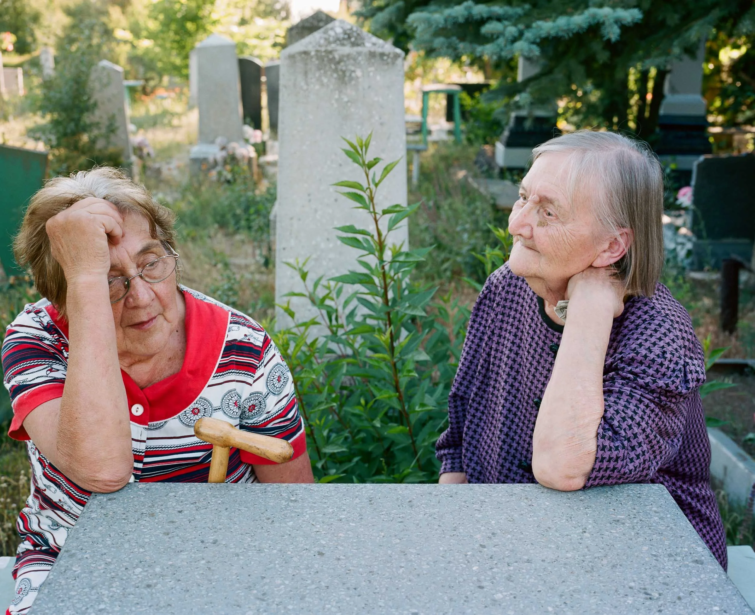 A documentary photograph of two elderly women sitting at a table outdoors in a cemetery, having a conversation. One woman wears a striped shirt with a red collar and glasses, and the other wears a purple patterned shirt, resting her chin on her hand.
