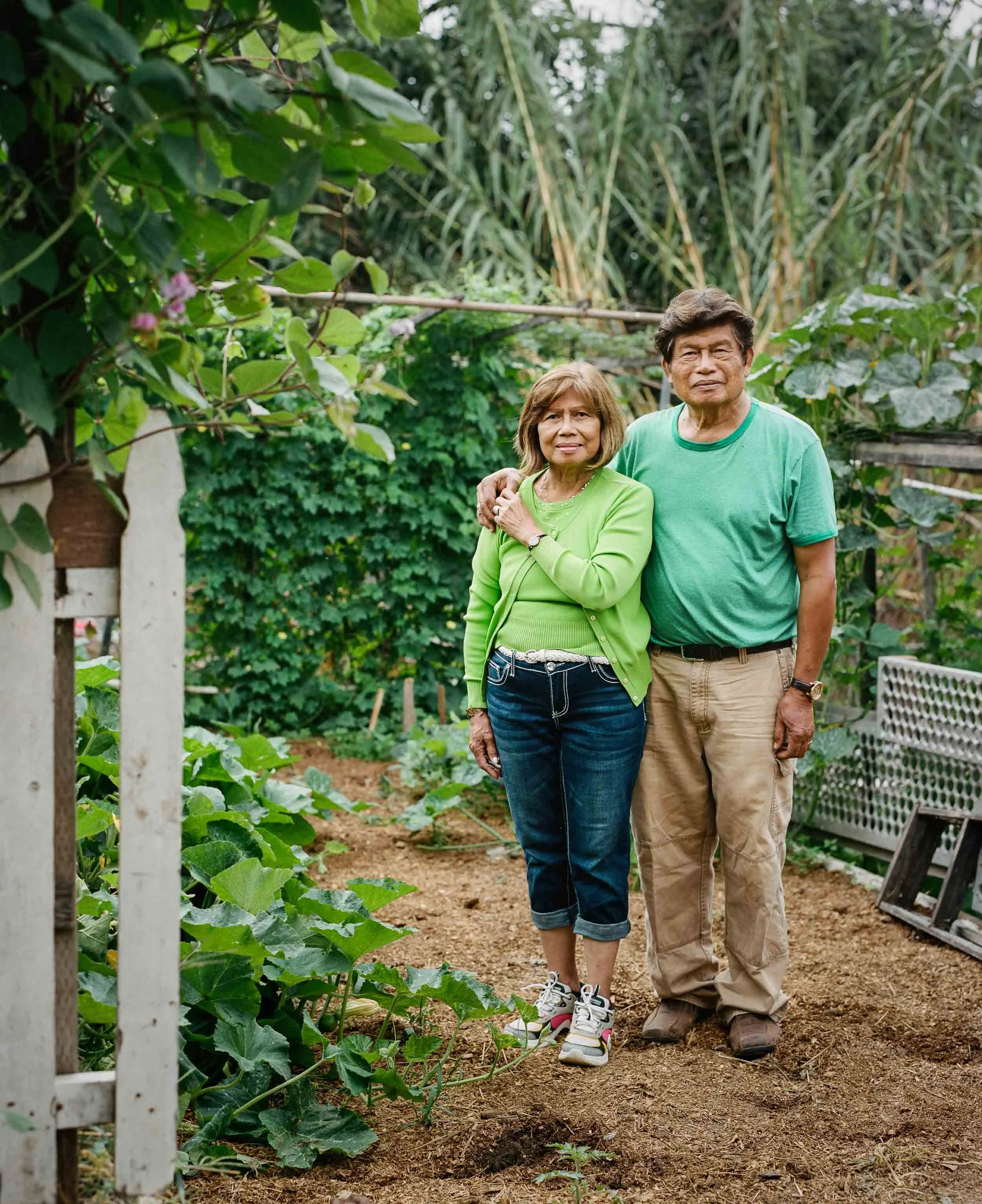 An elderly couple standing closely together in a lush garden with green plants and trees, dressed in green shirts and casual pants, smiling gently at the camera.