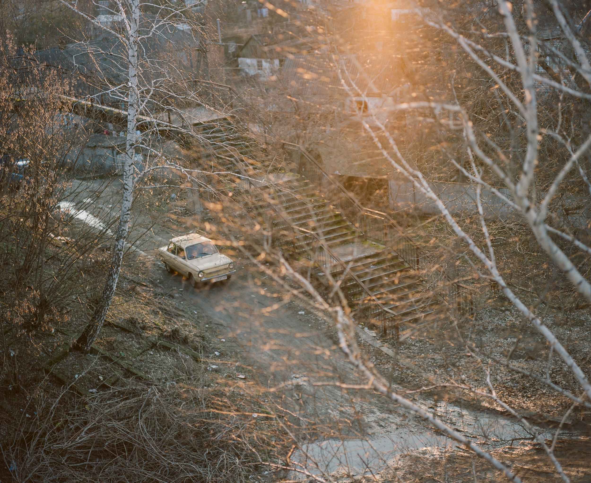 A landscape photograph with an old, beige car parked on a dirt pathway amidst leafless trees and a staircase leading up a hill, with sunlight filtering through the branches.