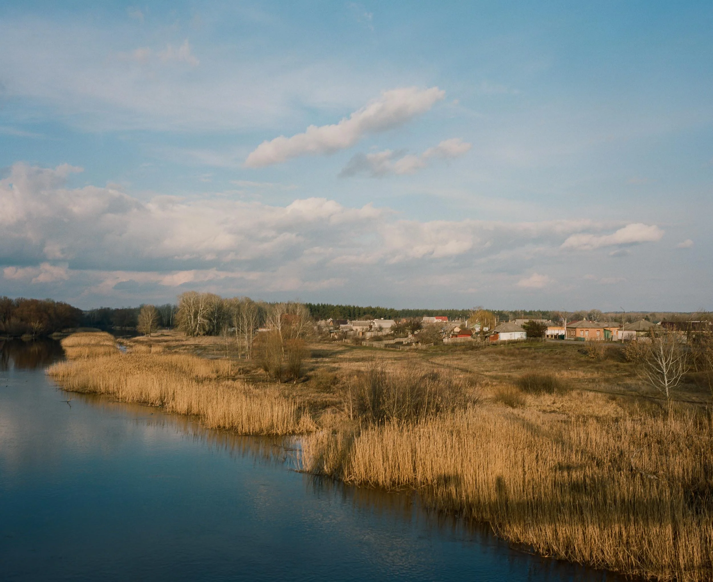 A photograph of a rural landscape with a river, grassy banks, trees, and a small village with houses under a partly cloudy sky.