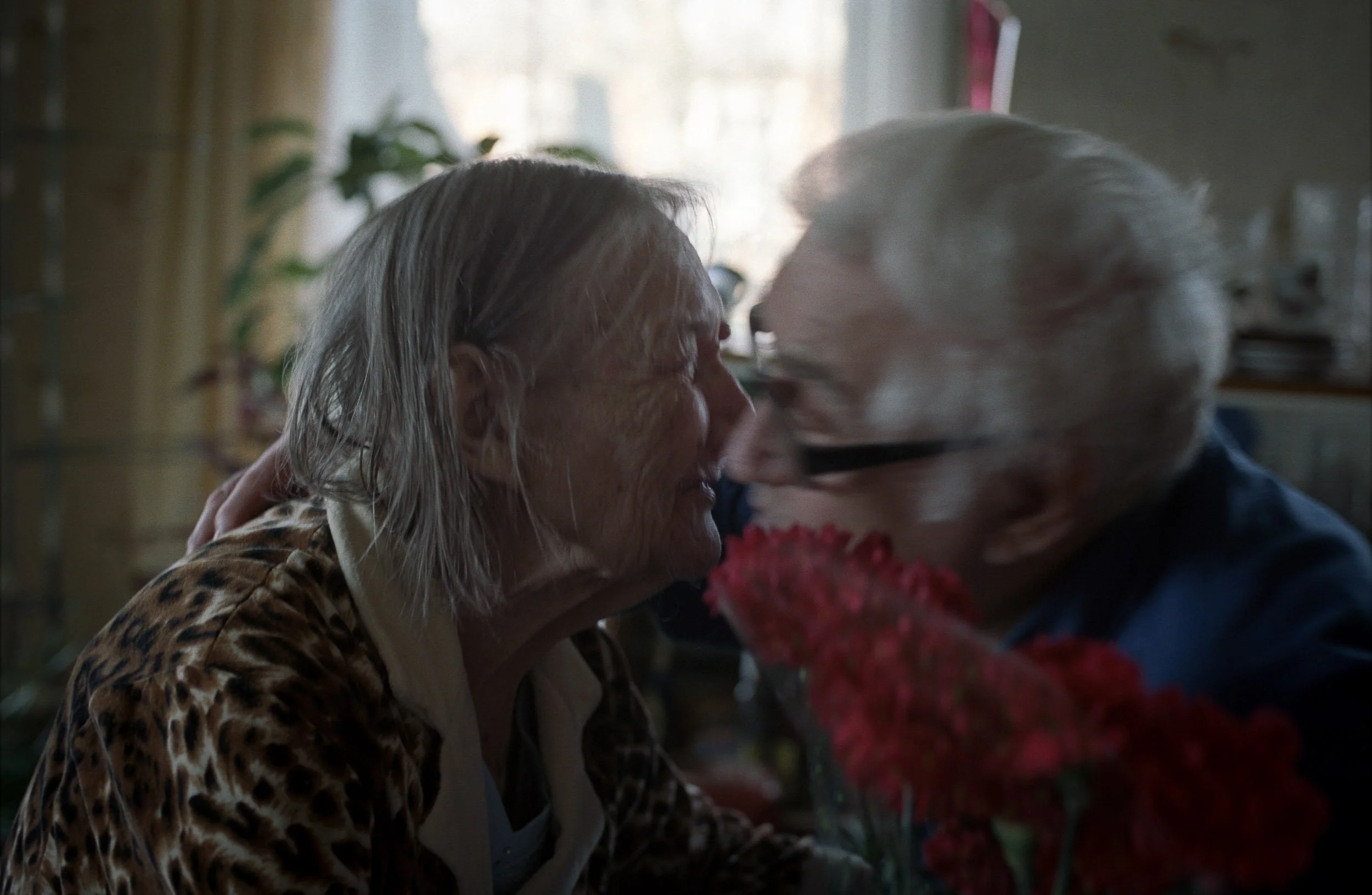 A documentary photograph of a senior woman and an elderly man share a close, affectionate moment indoors, with the woman smiling and the man gently leaning towards her, surrounded by home furnishings and a bright window in the background.