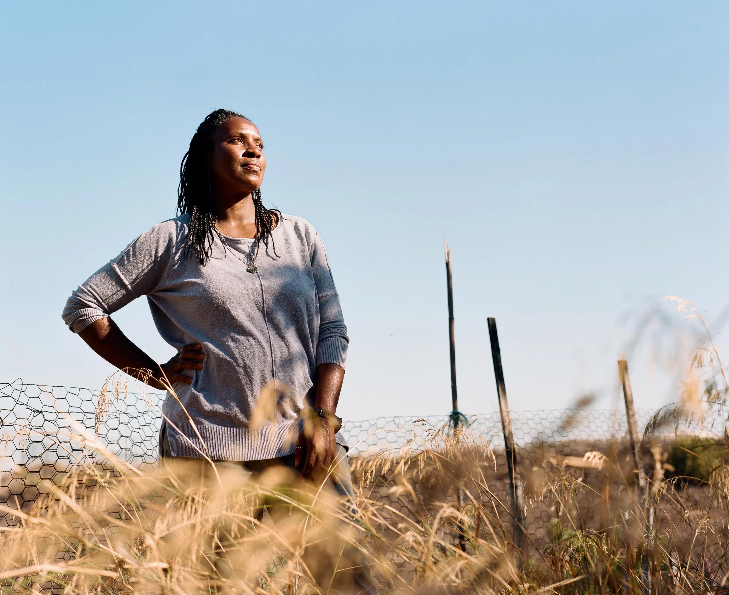 An environmental portrait of a woman standing in a field of tall grass, looking up towards the sky, with a fence in the background and a clear blue sky above.