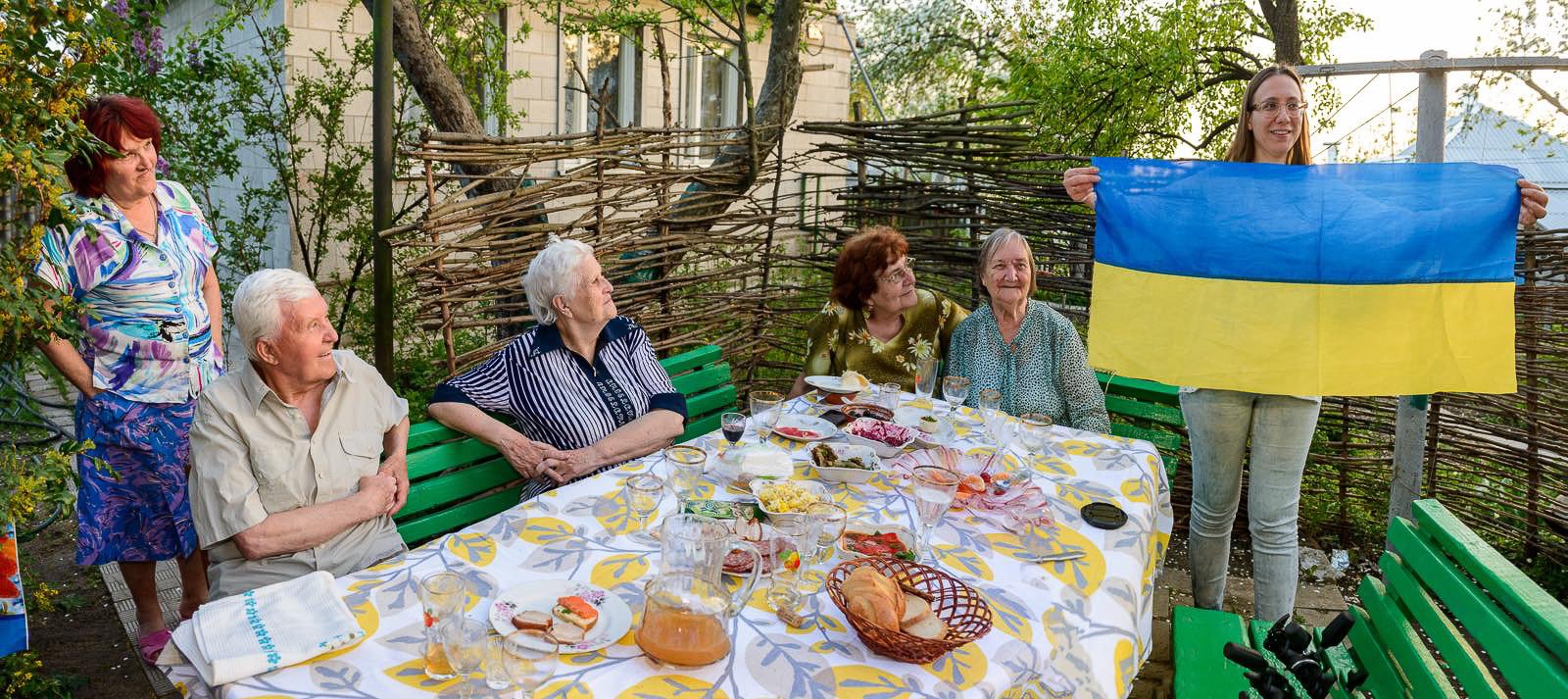 A photograph of a group of five women and one man gathered around a table outdoors, with one woman standing and holding a Ukrainian flag, celebrating together amid a garden setting.