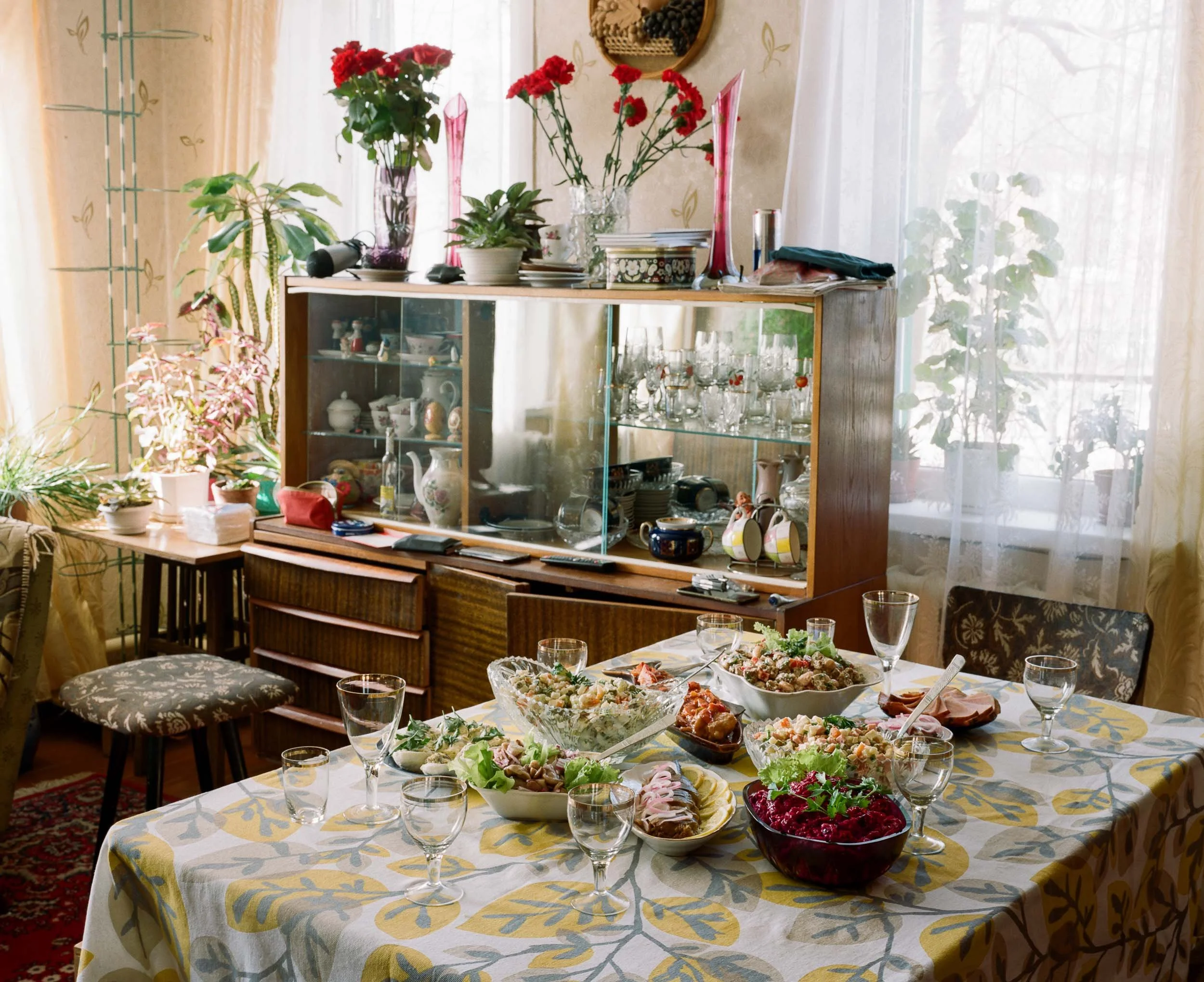 A dining room with a table set with various dishes, glasses, and utensils. The table has a floral tablecloth. There are multiple potted plants and decorative items on a sideboard and on the windowsill. The room is sunny with sheer curtains allowing natural light.