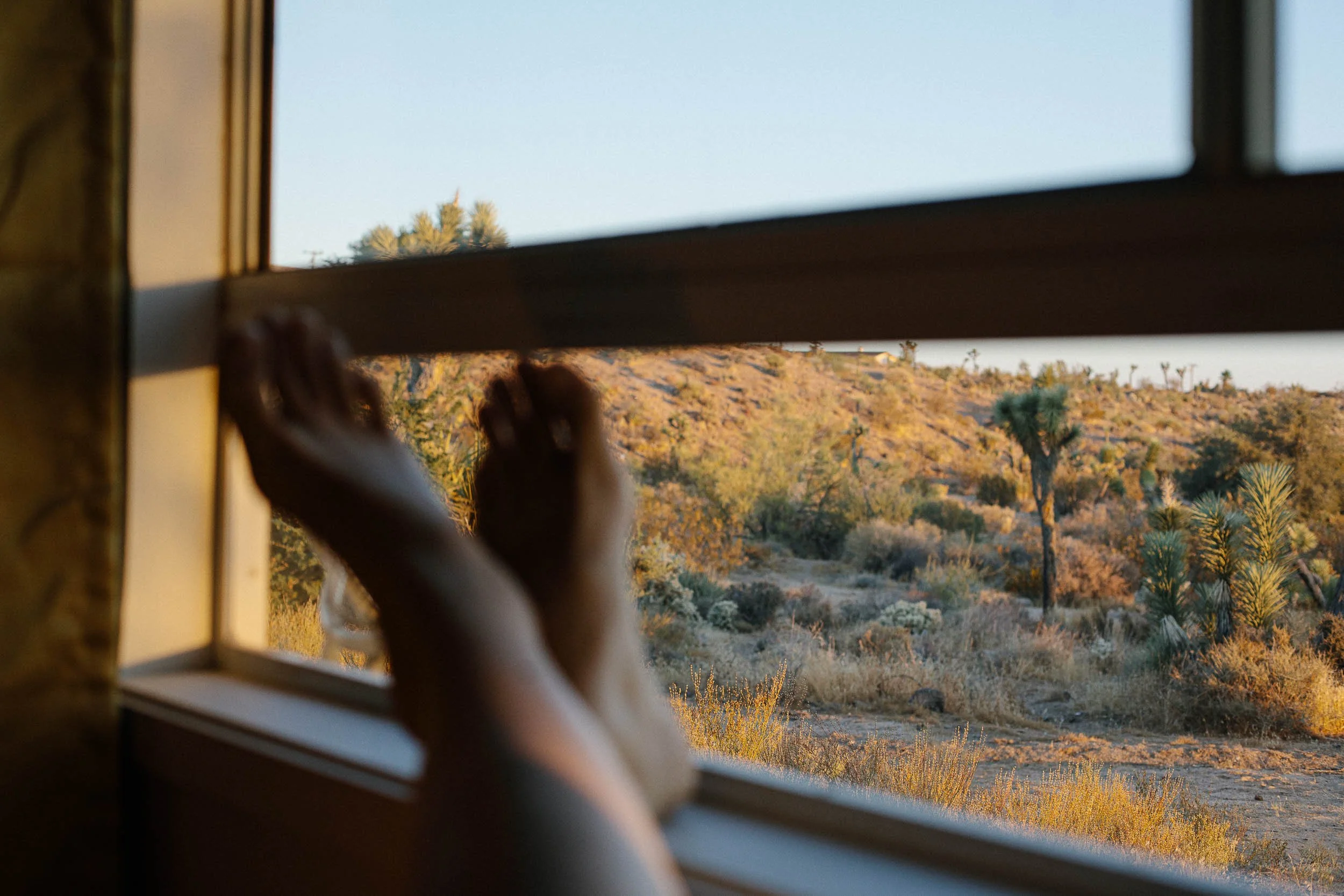 A commercial photograph of a person's bare feet resting on a windowsill, looking out at a desert landscape with trees and bushes during sunset.