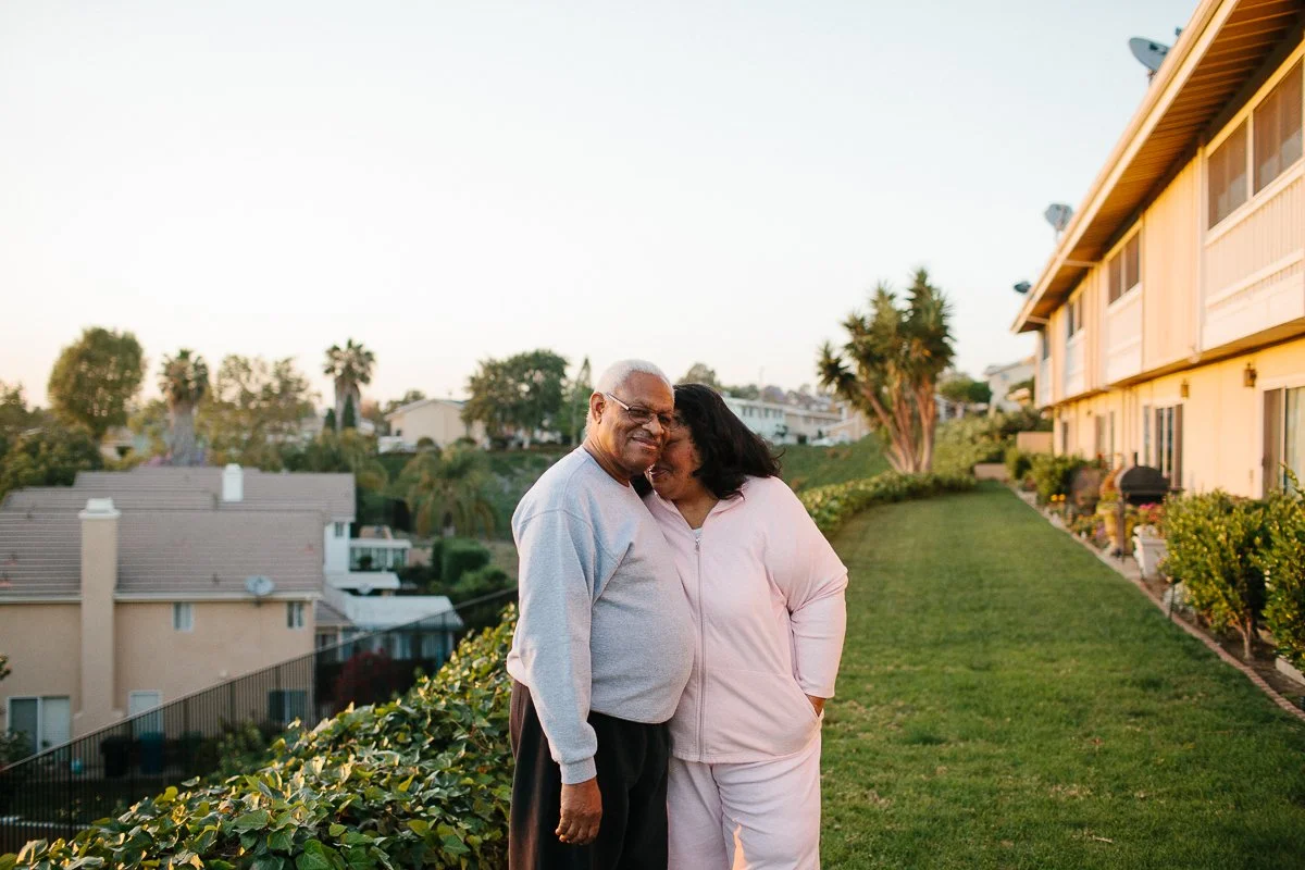 An environmental portrait of an elderly man and woman smiling and hugging on a grassy hillside with residential buildings and palm trees in the background during sunset.
