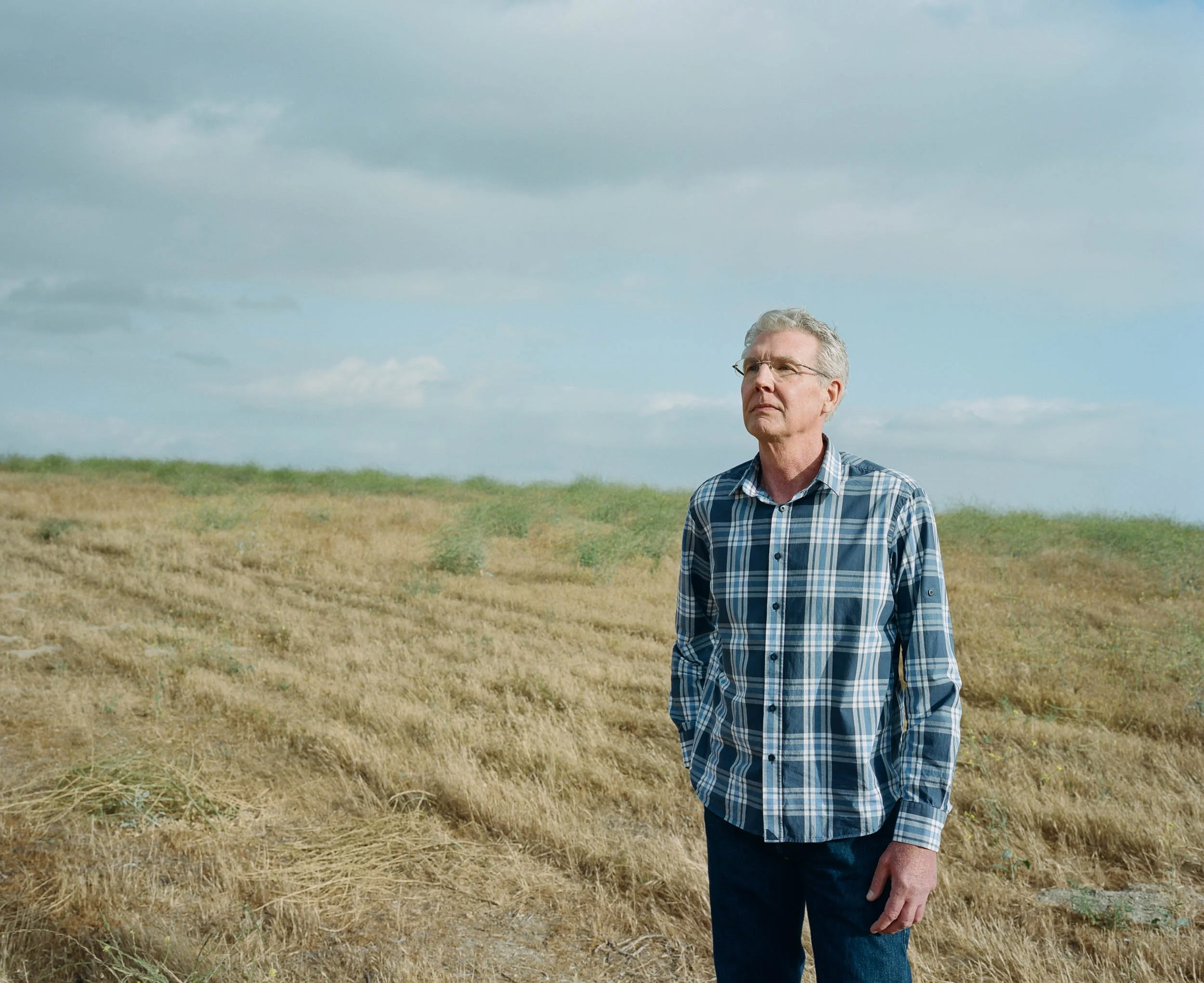 An environmental portrait of an elderly man standing alone in a field of dry grass under a cloudy sky, looking thoughtful.