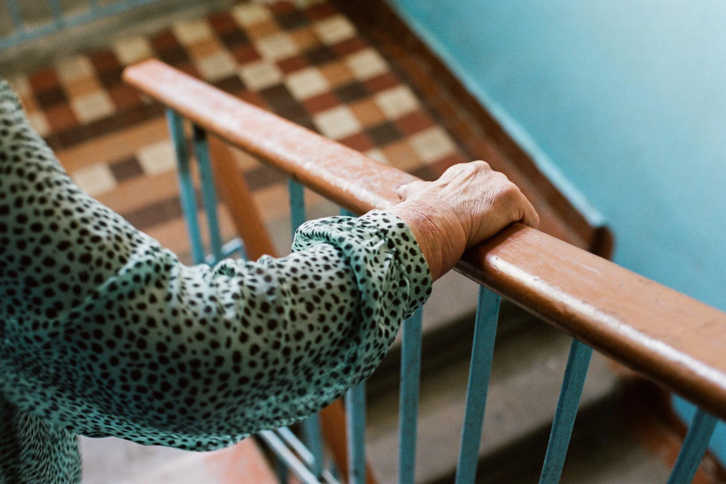 A close-up documentary photograph of an elderly person's hand gripping a wooden handrail on a staircase, with a patterned long-sleeve shirt.