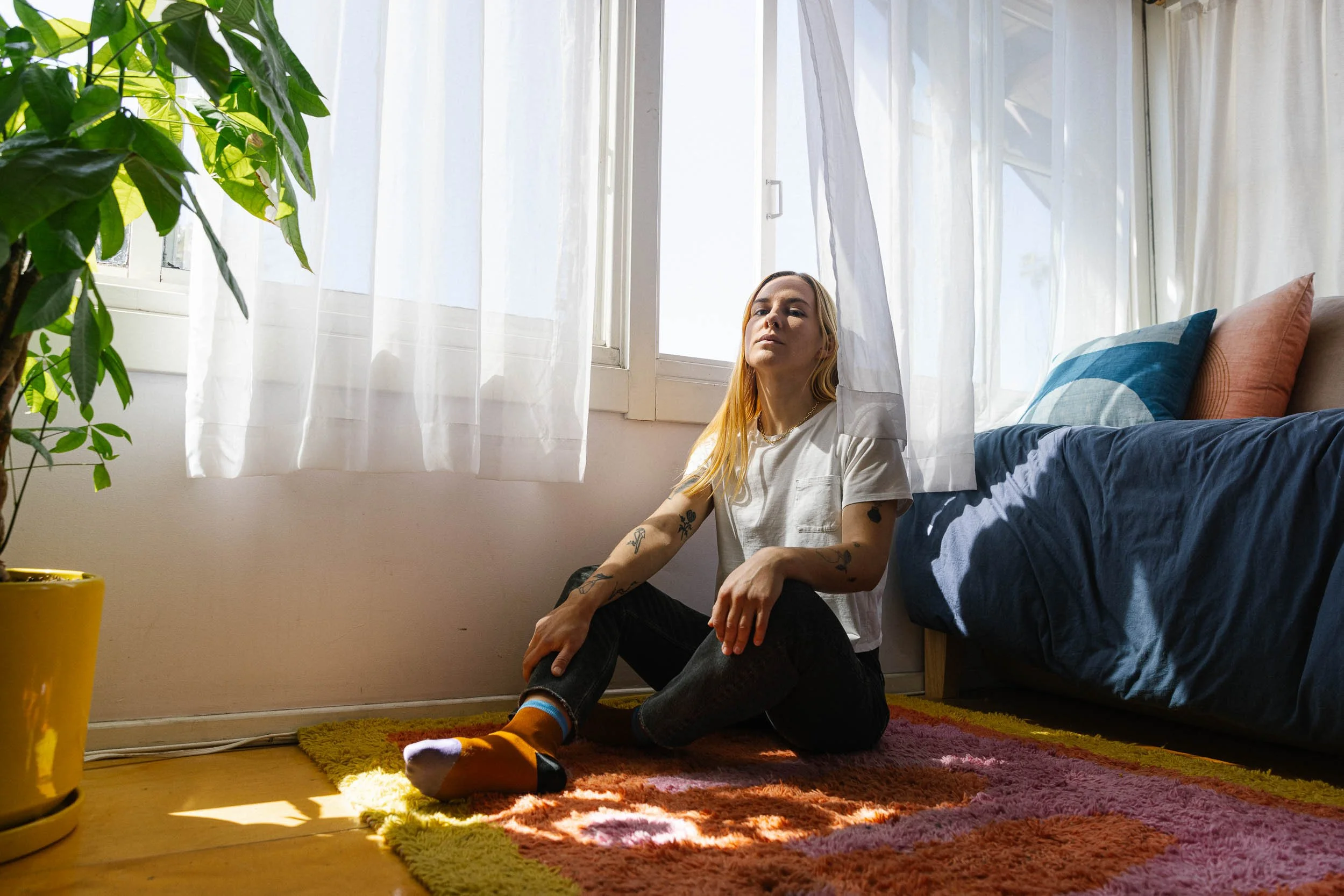 An environmental portrait of a young woman with long blonde hair sits on a colorful rug in a sunlit room, near a bed with blue and peach pillows.