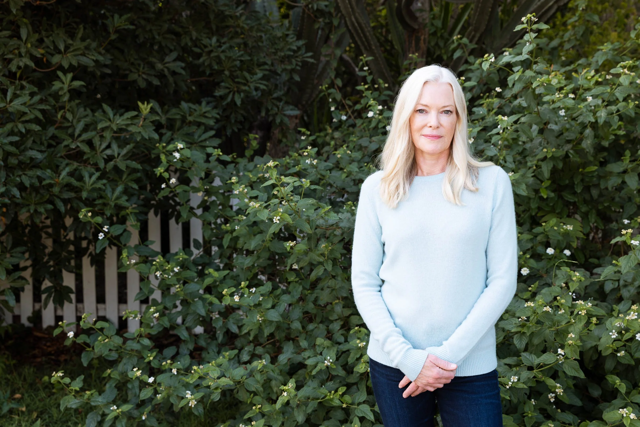 An environmental portrait of a woman with long blonde hair standing outdoors in front of green leafy bushes and white flowering plants, wearing a light blue sweater and dark blue jeans.