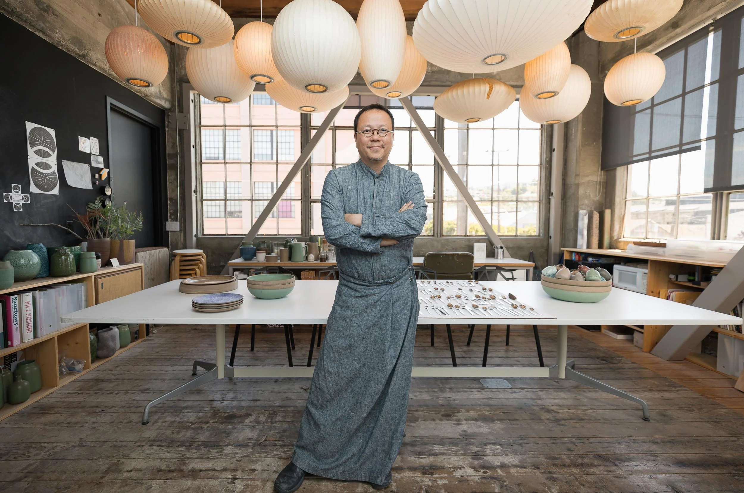 An editorial environmental portrait of a man standing in art studio with arms crossed, surrounded by pottery and jewelry displays, large lanterns hanging from ceiling, large windows in background.