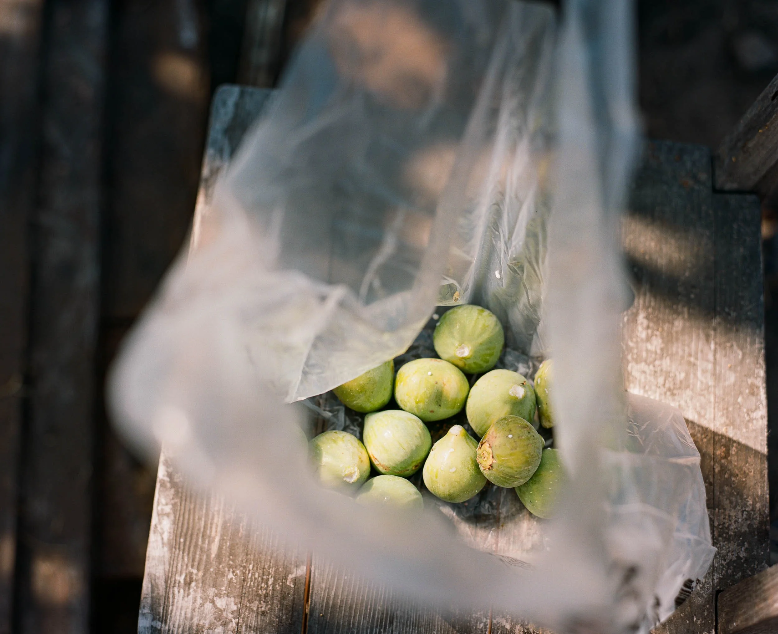 A still life detail photograph of green figs placed inside a transparent plastic bag, viewed from above on a rustic wooden surface.