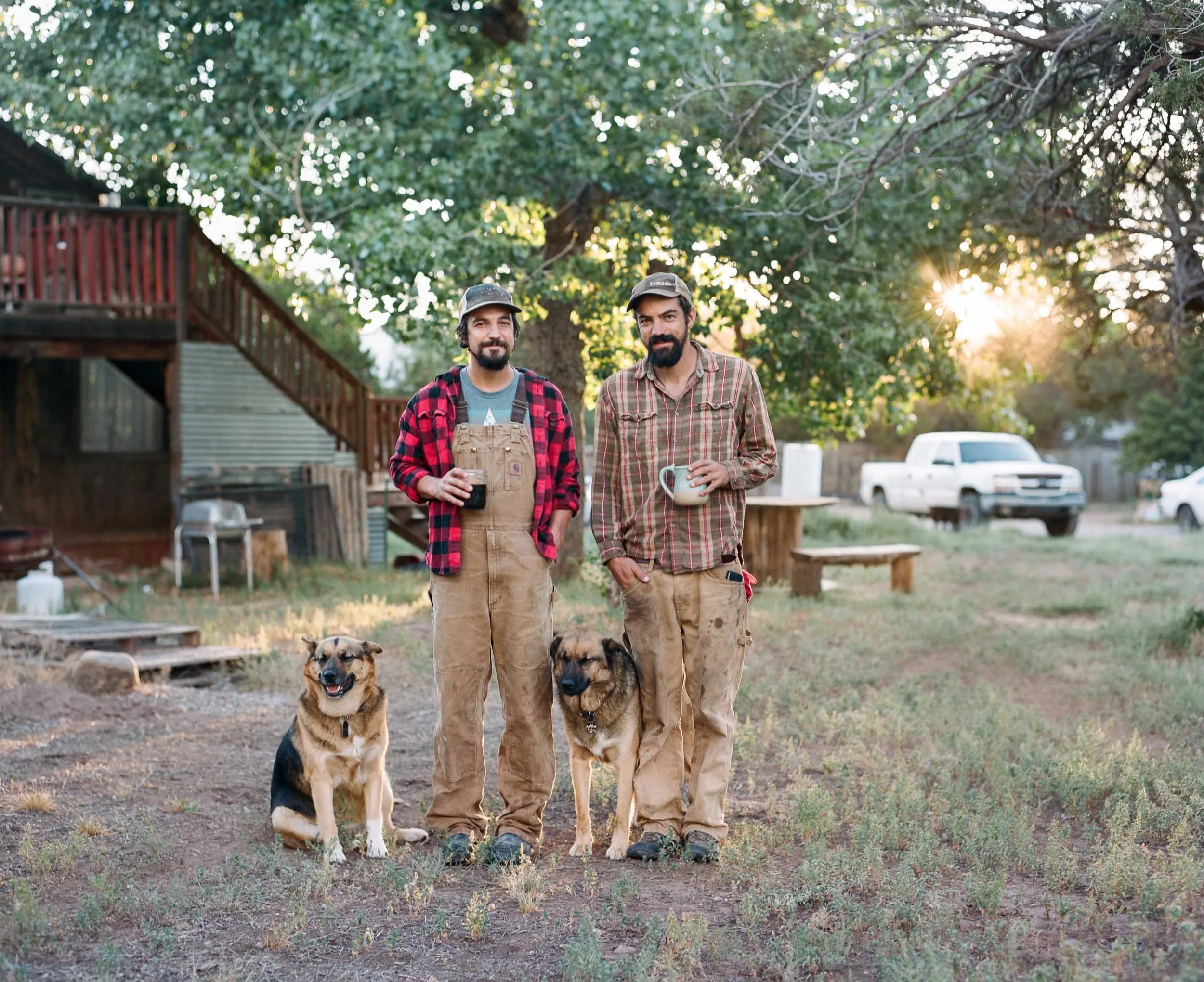 An documentary editorial environmental portrait of two men with beards and casual work clothes standing outdoors with two dogs, holding mugs, with a tree and a setting sun in the background.