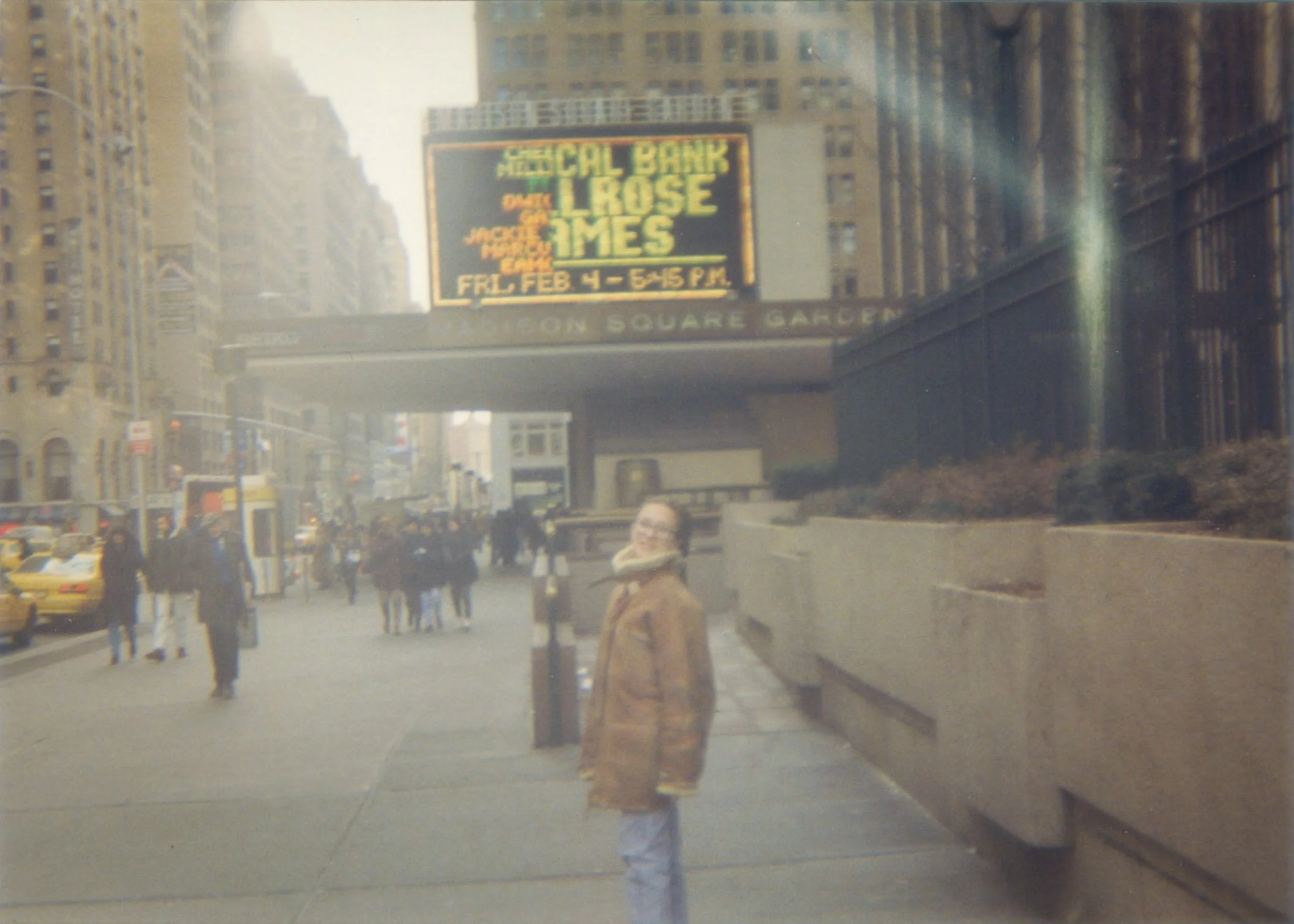 A young woman standing on a city sidewalk near a subway entrance, with tall buildings and a digital billboard advertising a concert, in New York City.