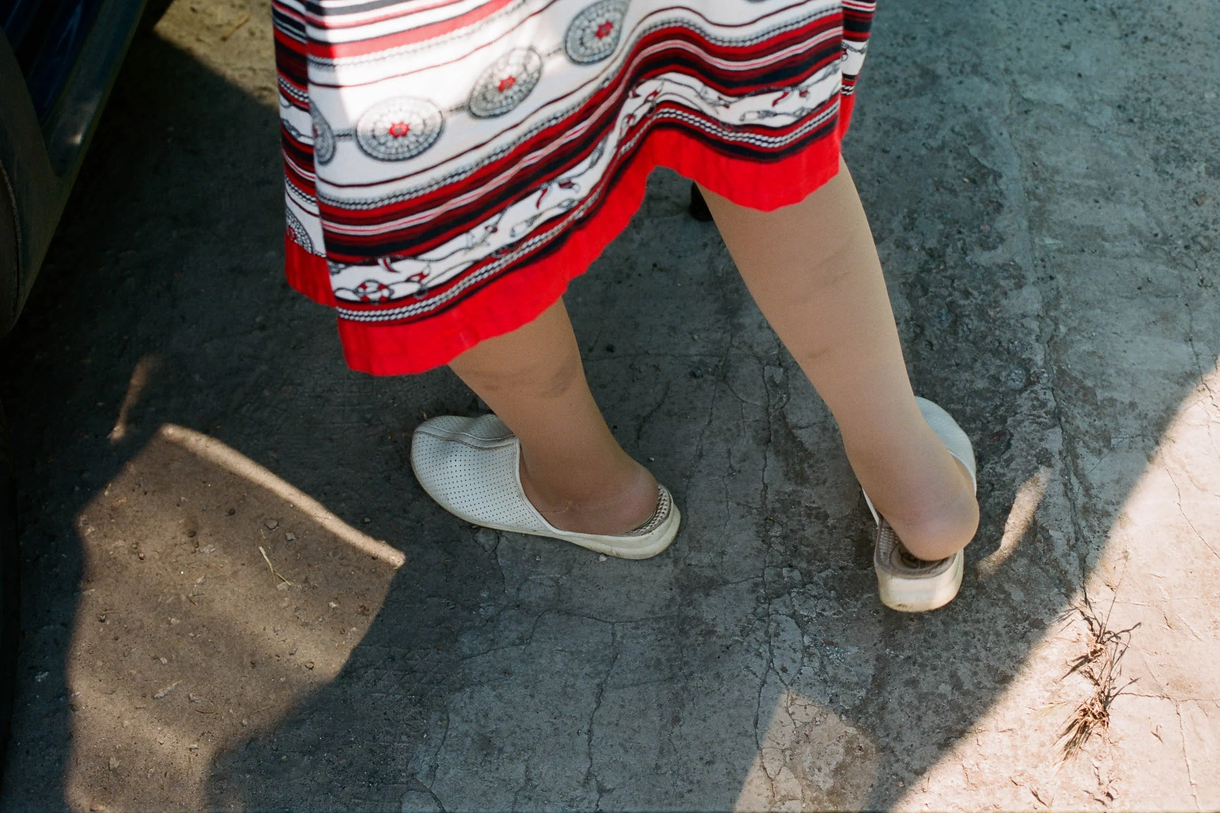 Person wearing a red, white, and black patterned skirt and white shoes, standing on cracked asphalt ground, casting a shadow.