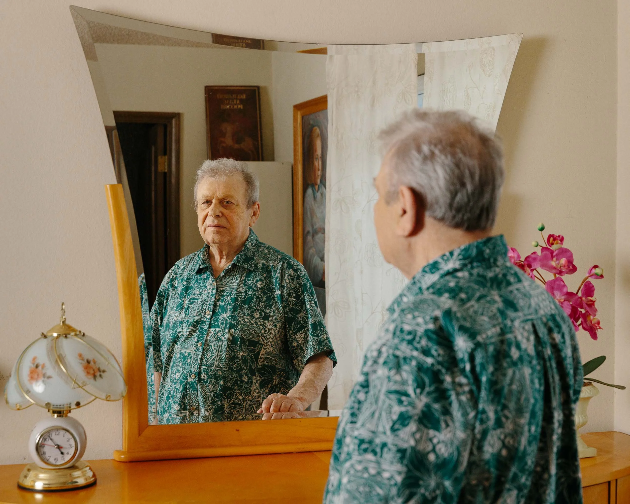 An editorial portrait of an elderly man with gray hair wearing a tropical patterned shirt looking into a mirror on a wooden dresser. A pink orchid is on the dresser, and a vintage clock with a white shade with floral design is next to it.