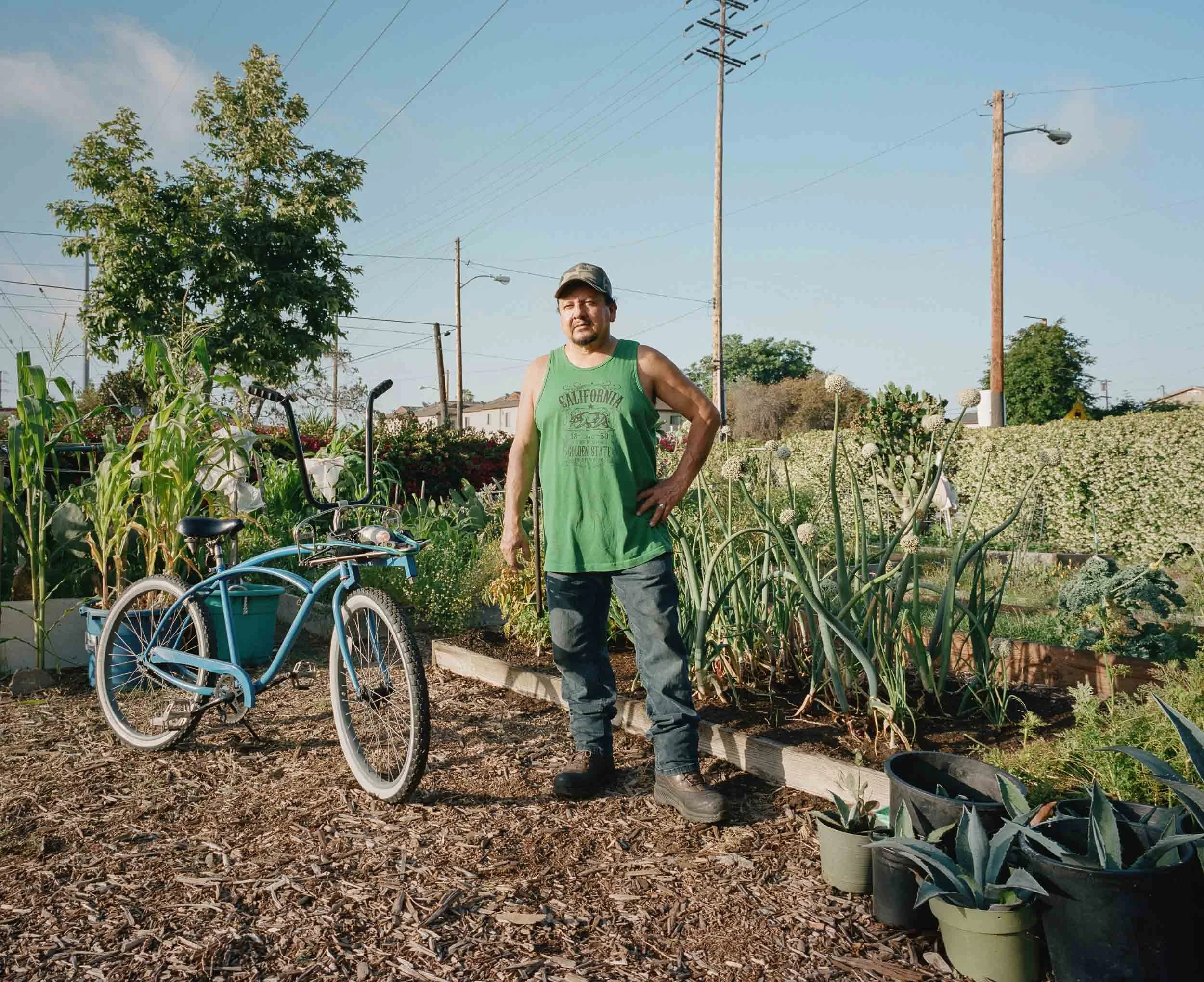 An environmental portrait of a man standing in his garden next to a blue bicycle, wearing a green tank top and dark jeans, with various plants and flowers around him and power lines in the background.
