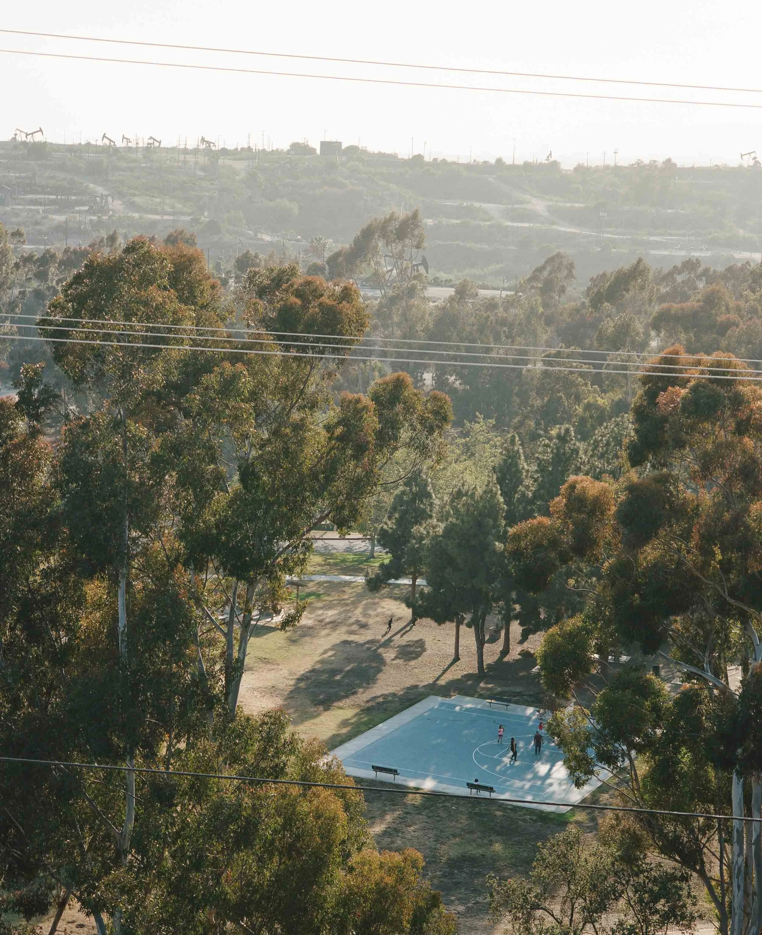 A landscape photograph of an outdoor basketball court surrounded by trees with a few people playing. In the background, there is a landscape with hills and oil pumps.