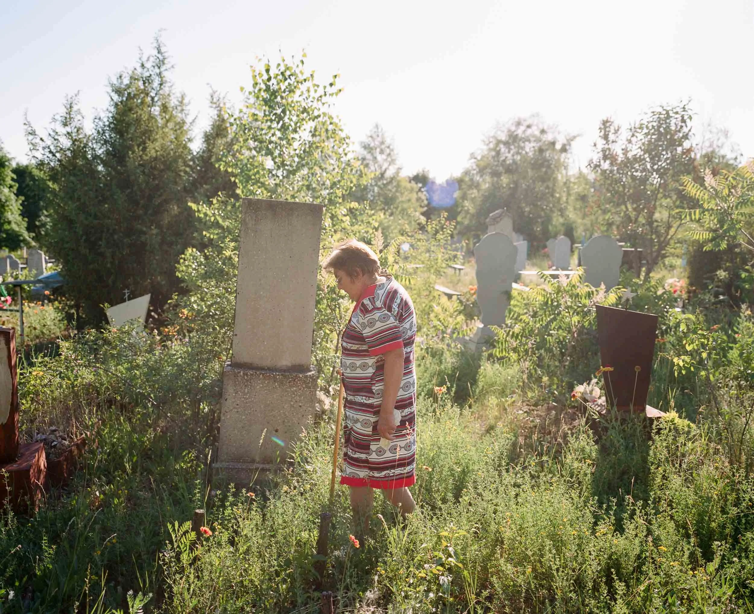 A documentary photograph elderly woman in a patterned dress stands in a grassy graveyard, looking at a tombstone.