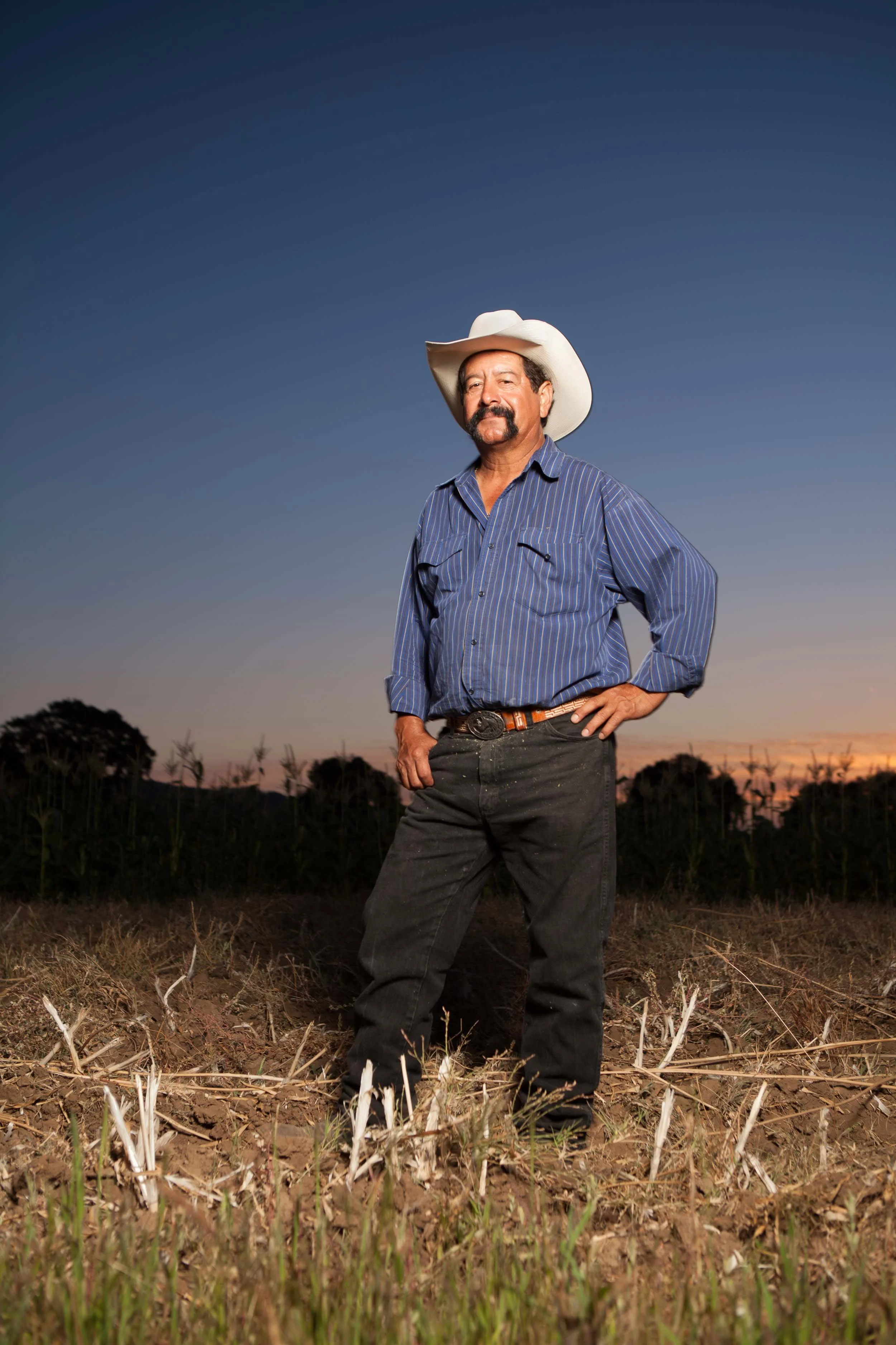 An environmental editorial portrait man standing in a field at sunset, wearing a cowboy hat, blue striped shirt, and dark jeans.