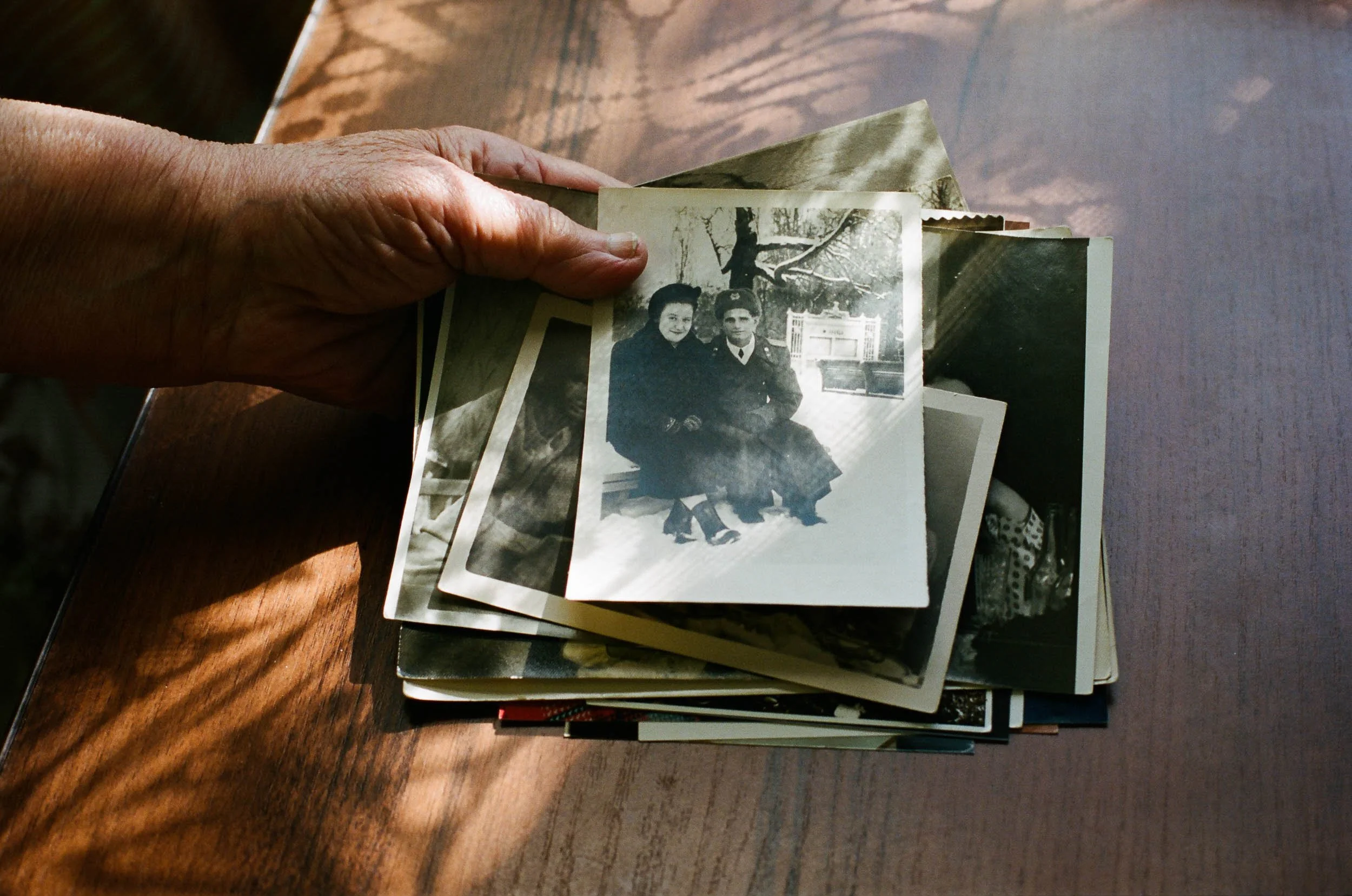 A documentary photograph of a person holding a black-and-white photograph of two women sitting outdoors on a wooden bench, with a tree and a fence in the background.
