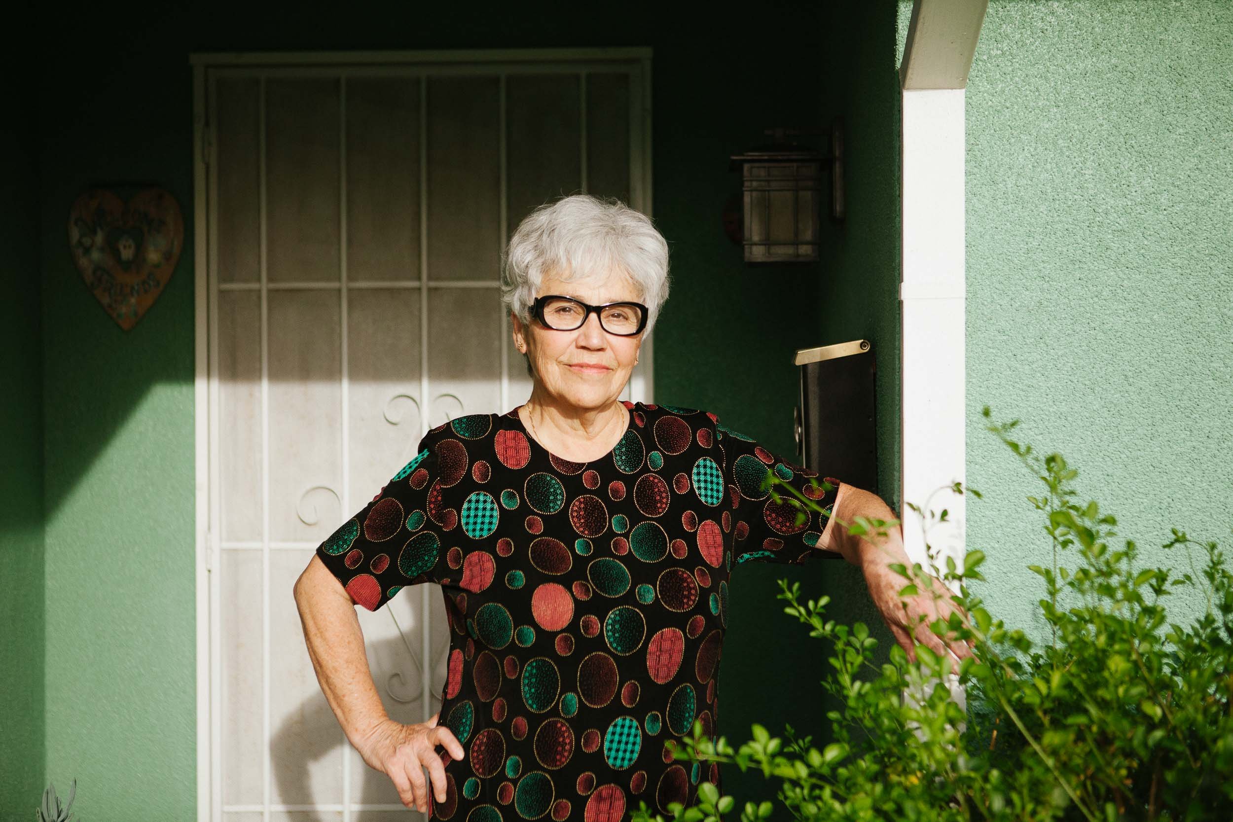 An elderly woman with short gray hair, black glasses, wearing a black dress with multicolored circular patterns, standing at a home's doorway with a green wall, white door, and some greenery in front.